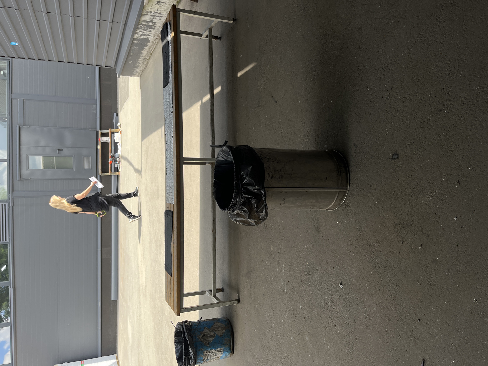 A blonde woman in a black t-shirt and black pants walks from left to right across a concrete outdoor area. She is holding a white paper in her right hand and a green object in her left. A long, wooden bench with metal legs is situated in the middle ground of the image, with two trash cans on either side of it. The trash can on the left is blue and appears to be made of plastic, while the trash can on the right is metallic and cylindrical. The woman is walking towards the right side of the image, and the bench and trash cans are positioned in front of a gray building with a large door. The scene appears to be an outdoor seating or waiting area. The location is not specified to be in Kyiv, Ukraine.