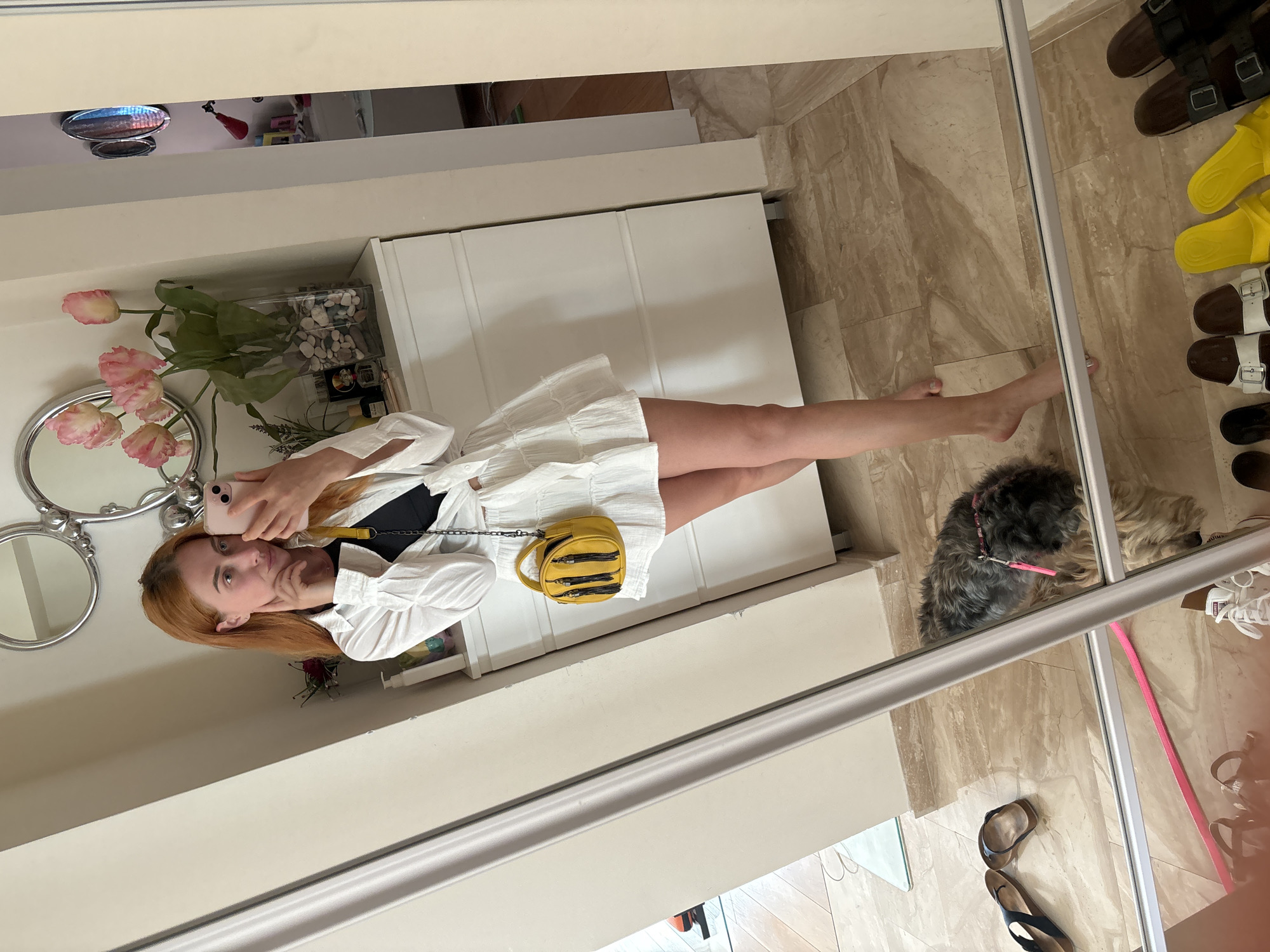 A young woman with long, auburn hair is taking a mirror selfie in what appears to be a well-lit room. She is wearing a white shirt dress with ruffled detailing and a black bra or bralette underneath. A small, yellow crossbody bag with multiple zippers hangs from her shoulder. She is holding a pink smartphone in her right hand, capturing the reflection.
In the background, a large white closet or cabinet is visible. To the left, there are decorative mirrors with ornate silver frames, and a vase filled with pink flowers and greenery. To the right of the closet, on the tiled floor, is a small dog with grey and tan fur, wearing a pink collar and leash. Several pairs of sandals and flip-flops are also visible on the floor, reflecting in the mirror. The floor is tiled with a beige and brown marble pattern.
The overall impression is of a casual moment, possibly in a home or dressing room, with the sunlight suggesting daytime. The setting appears tidy and organized, with decorative elements that add a personal touch. The woman's expression is relaxed and perhaps a little playful as she poses for the picture. The environment is indoors, with clear lighting and no indication of weather conditions. The presence of the dog suggests a domestic setting.