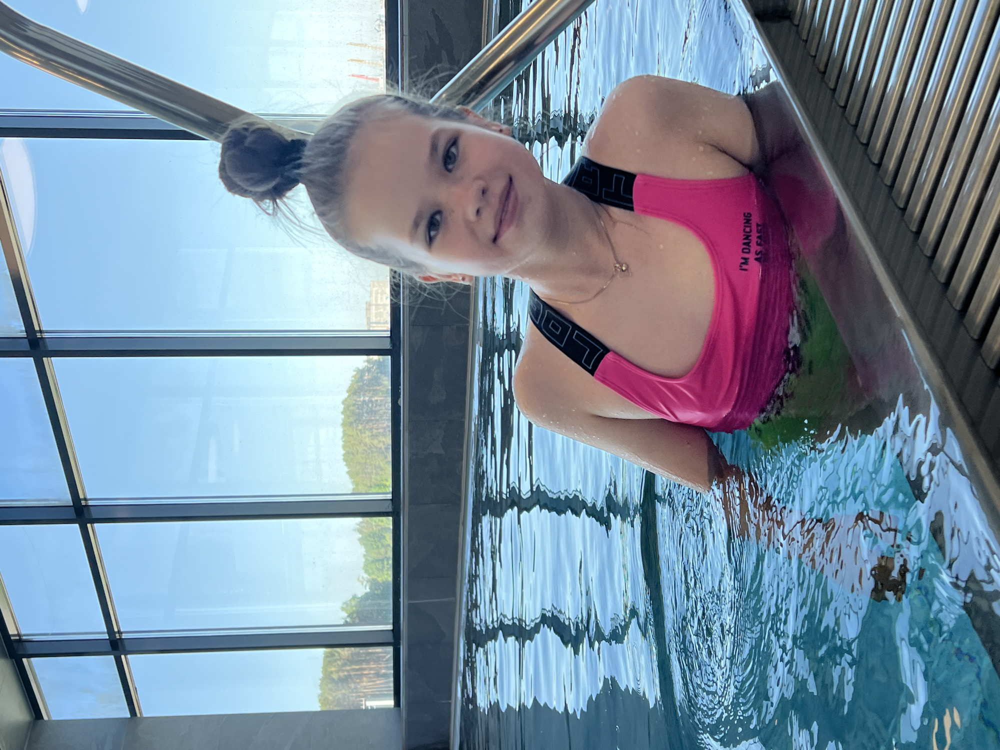 A young girl is partially submerged in a pool, wearing a pink swimsuit with the words "I'M DANCING AS FAST" printed on the front. She has her hair in a bun and is smiling towards the camera. The pool is located indoors, near a large window with a view of trees. The indoor pool is located in the city of Chaikovsky, Russia. A metal bar runs along the side of the pool.