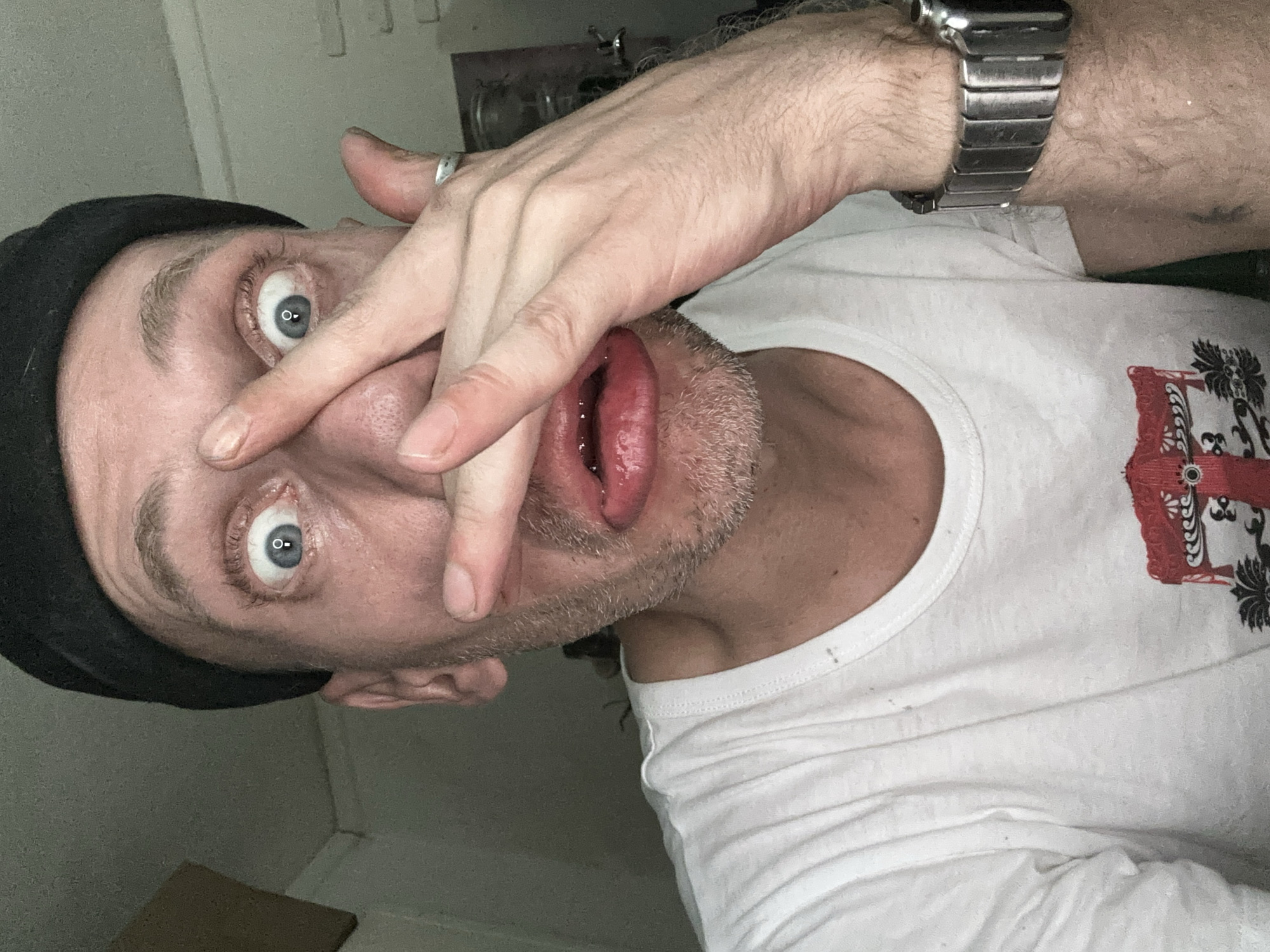 A close-up, self-portrait photograph shows a Caucasian man wearing a black beanie and a white t-shirt with a graphic print. His face is partially obscured by his hands, which are positioned over his eyes and mouth. His eyes are wide open, with one hand pressing on the bridge of his nose and the other across his lips. He has stubble on his chin and a ring on his left ring finger. A metal watch is visible on his right wrist. The background is dimly lit and appears to be an indoor setting with a textured wall and some indistinct objects. There is no identifiable location in the city of Moscow, Russia, visible in this image.