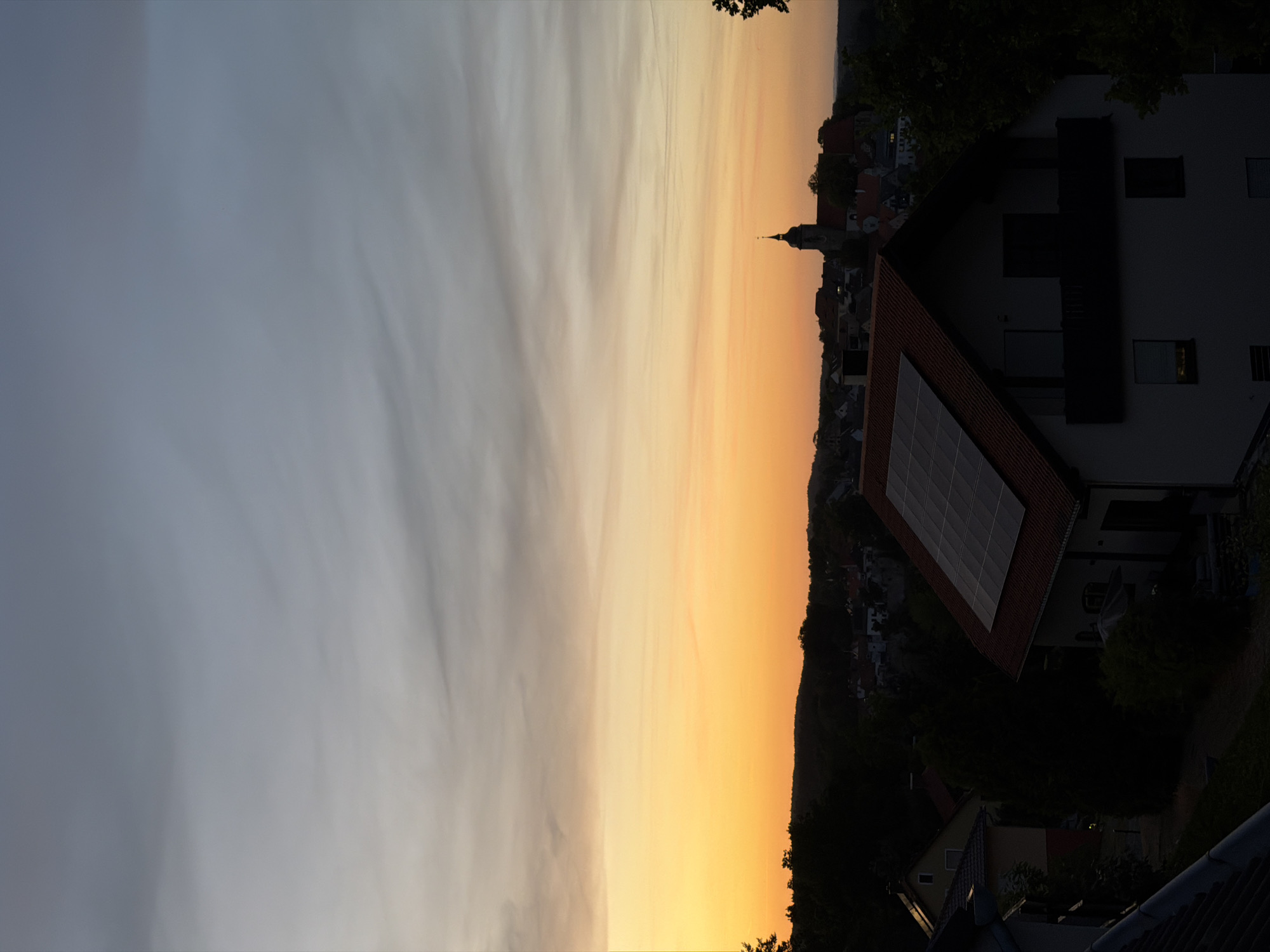 The image shows a view over rooftops in the city of Creußen, Germany, during a sunset. The sky displays a gradient of colors, transitioning from a pale blue at the top to a vibrant orange at the horizon. In the foreground, a house with a red-tiled roof and solar panels is visible. Further in the background, a church steeple punctuates the skyline, surrounded by other buildings and trees. The overall impression is a peaceful, residential scene at dusk.