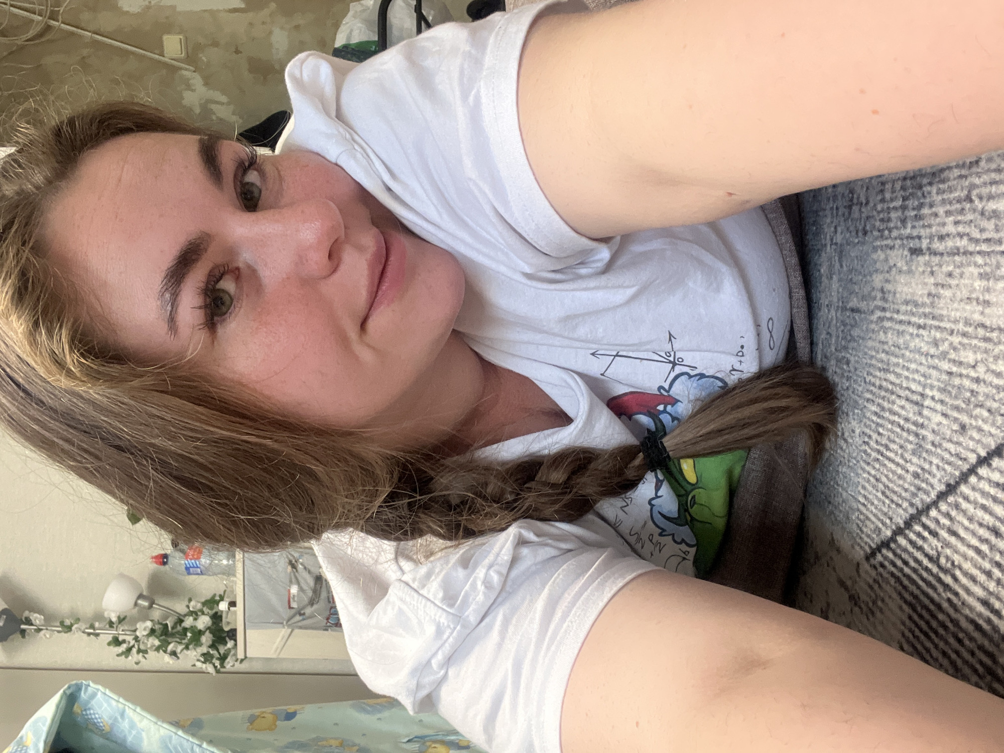 A woman with light brown hair styled in a braid is pictured in a close-up, angled selfie. She wears a white T-shirt. A patterned, gray and white blanket or sofa is visible behind her. In the background, a lamp and a shelf with various items are partially visible. There is no indication that this photograph was taken in the city of Chaikovsky, Russia.
