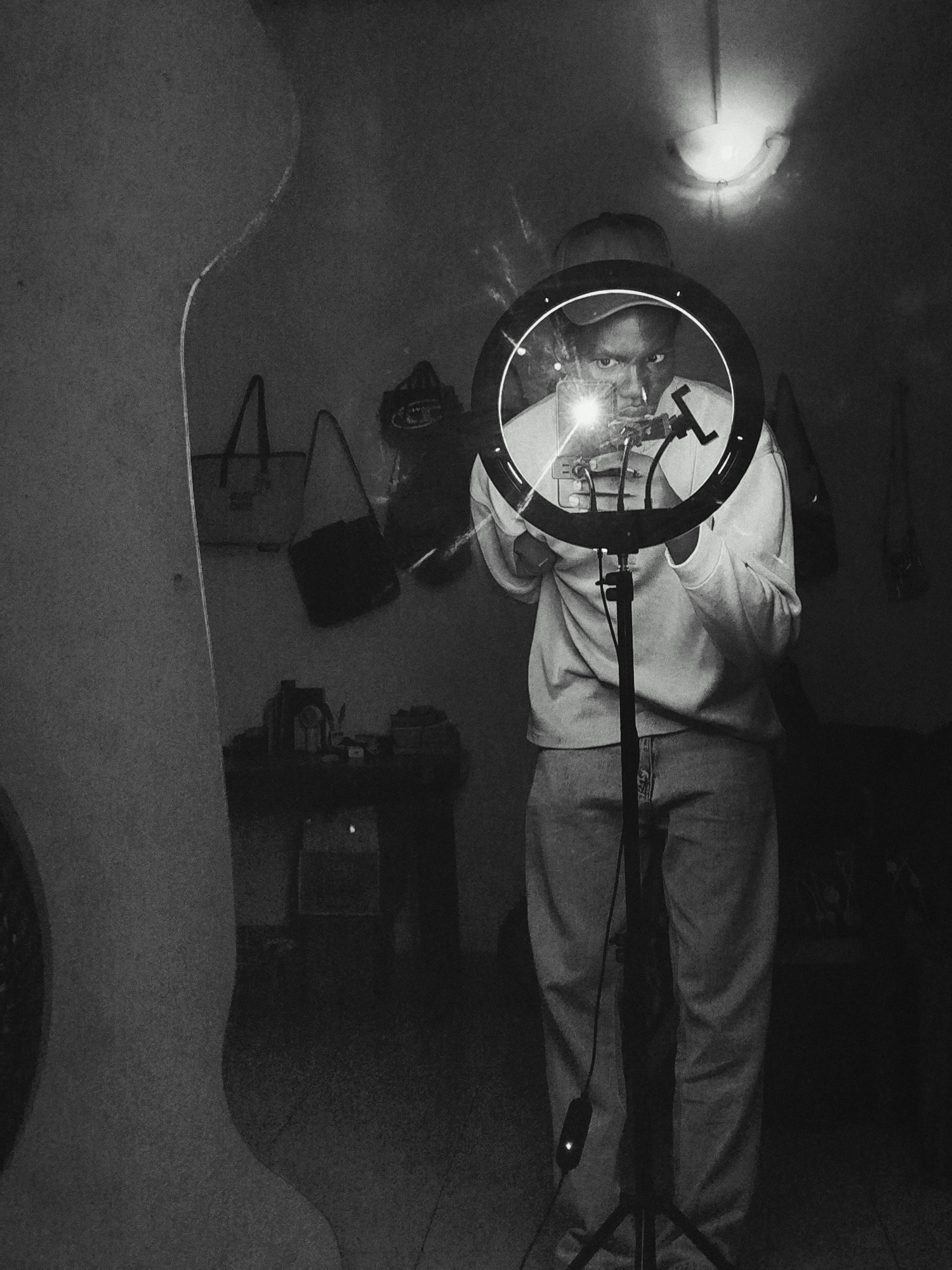 The image is a monochrome indoor photograph featuring a person standing in front of a ring light, presumably taking a selfie or recording a video. The person is wearing a baseball cap, a light-colored sweatshirt, and jeans. They are holding a smartphone in their hands within the center of the ring light. The room appears to be cluttered with various items, including bags hanging on the wall, a table with objects on it, and an overhead light fixture. The location is Unknown, Unknown.