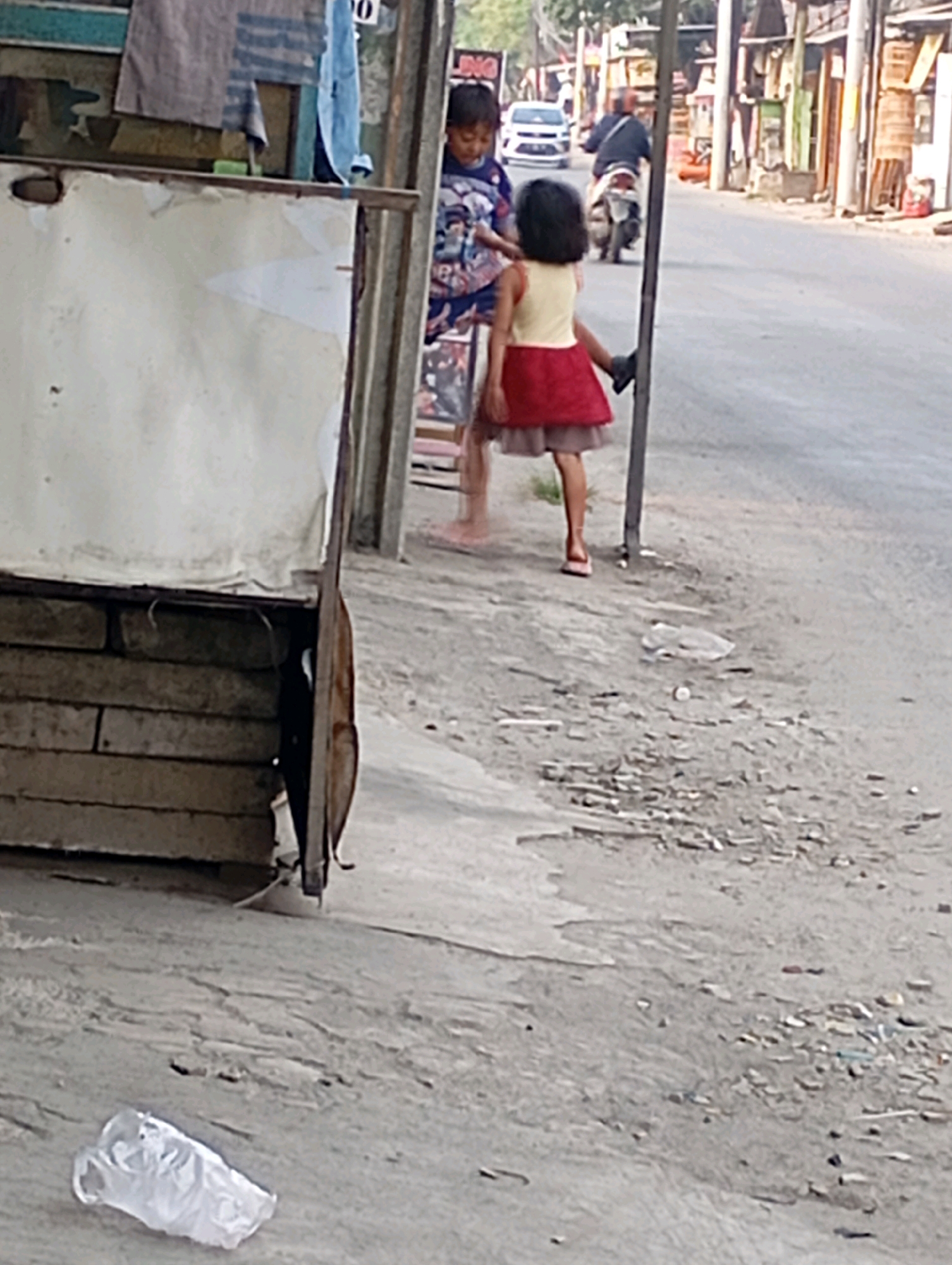 The image depicts a street scene in Babelan, Indonesia. In the foreground, a weathered wooden structure with a white tarp is visible. Two young girls are standing on the pavement. The girl on the right wears a red and yellow dress, while the girl on the left wears a blue shirt with a patterned design. Behind them, a white car and a person on a motorcycle are visible on the paved road. The environment appears to be an urban or semi-urban setting with buildings lining the street and debris scattered on the ground. A discarded plastic bag lies on the pavement in the lower left corner of the image.