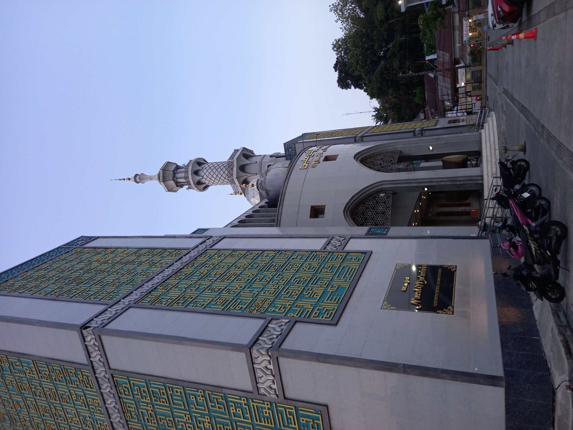 The image shows the Masjid Nurul Mu'allimin in Samarinda, Indonesia, during the day. The mosque is a large, modern building with a prominent minaret. The facade of the mosque is decorated with intricate geometric patterns in gold and teal. In the foreground, several motorcycles are parked, and a car is visible in the background. The sky is clear and blue. The overall impression is one of a well-maintained and important religious structure.