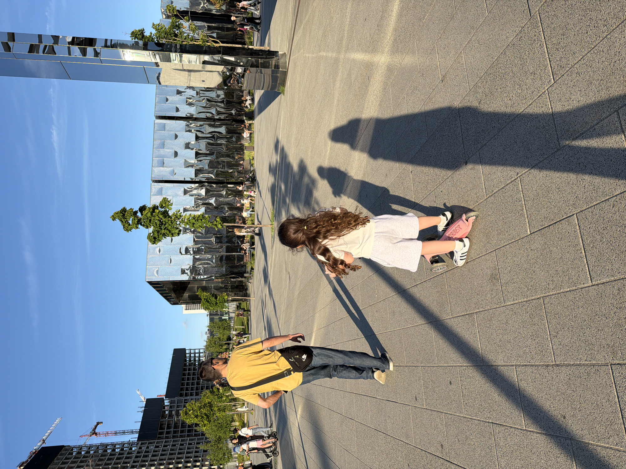 A man in a yellow t-shirt and blue jeans walks away from the camera, his arm at his side, a dark crossbody bag slung over his shoulder. A young girl with long, curly brown hair, wearing a white t-shirt and white shorts, rides a pink scooter in front of him. She is wearing white socks and black-and-white athletic shoes. They are on a paved plaza with large, square gray tiles. To the right, a tall, mirrored sculpture reflects the blue sky and surrounding buildings. In the background, to the left, are several unfinished high-rise buildings with cranes. To the right, a modern building with a mirrored facade is visible, with people seated on benches in front of it. The sun is low in the sky, casting long shadows across the plaza. The environment appears to be a public outdoor space in a city.