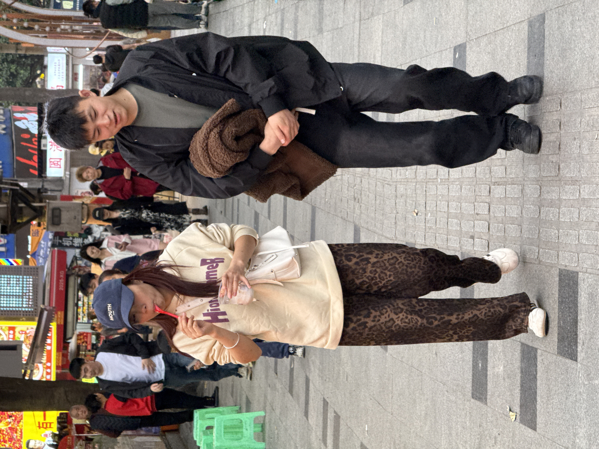 The image captures a street scene in Liangjiang, China. A young woman is eating with a spoon from a clear plastic container. She wears a beige sweater with purple writing on the sleeve, patterned pants, a blue baseball cap with the word "MOUTH," and a white purse over her shoulder. Standing next to her is a young man in a black jacket and black pants. He is holding a brown fabric item. The setting is a busy street with many people in the background. Colorful shopfronts and signage are visible. The street is paved with gray concrete tiles. Green plastic stools are stacked on the ground.