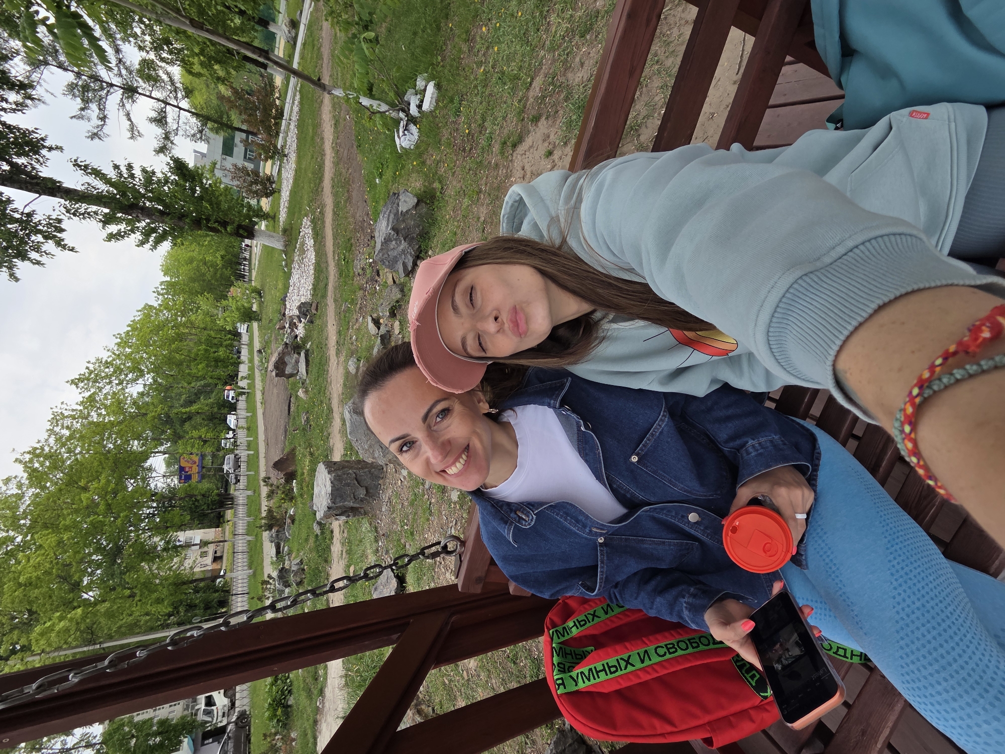 Two women are posing for a selfie on a wooden bench in what appears to be a park setting. The woman in the foreground is smiling at the camera while wearing a denim jacket over a white shirt, and is holding a red cup in one hand and a smartphone in the other. Next to her is a red backpack with green straps. The other woman, wearing a light blue hoodie and a pink cap, is making a kissing face at the camera. They are sitting on a wooden bench with metal chains visible on the side. The background includes grass, trees, and a few buildings. The location is Birobidzhan, Russia, judging from the Cyrillic text on the backpack, which translates to "smart and free."