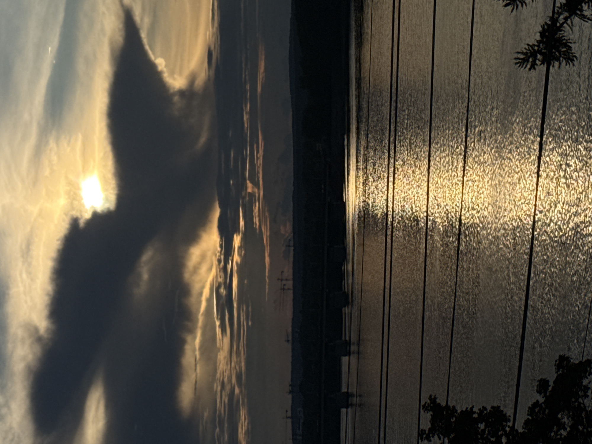 The image depicts a sunset over the Kama River in Perm, Russia. A large, dark cloud formation dominates the upper half of the frame, with the sun peeking through a gap, casting a golden glow on the sky. In the middle ground, a silhouetted bridge spans the river, with construction cranes visible on the distant shoreline. The lower half of the image shows the rippling surface of the river, reflecting the sunset's light. Several power lines are strung across the foreground, partially obscuring the view of the river and the distant landscape. Silhouetted trees frame the left and right edges of the image.
