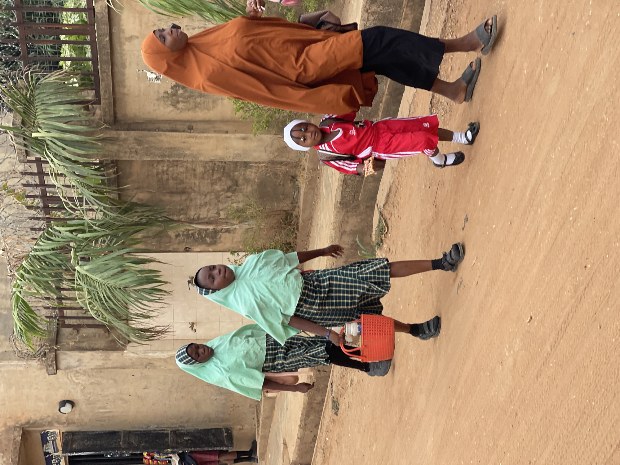 The image shows a street scene in Kuchingoro, Nigeria. Two schoolgirls in green headscarves and plaid skirts carry an orange plastic basket. Nearby, a woman in an orange hijab walks with a child in a red tracksuit. The street is unpaved. A building with a metal fence and palm trees is in the background.