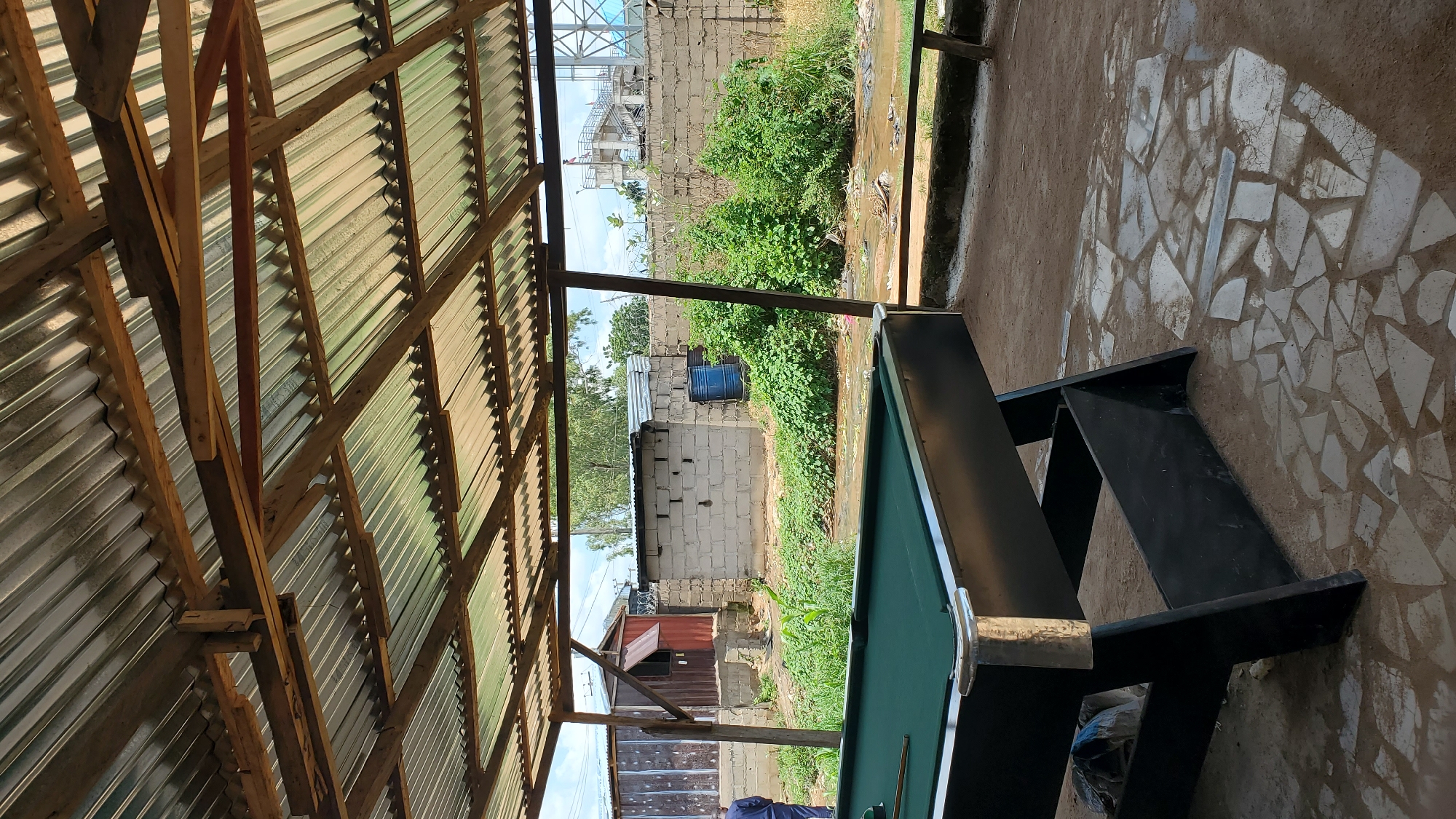 A full shot of a covered outdoor area with a pool table in the foreground, located in Jalingo, Nigeria. The pool table is dark green with black legs and silver accents. Behind the table, there is a dirt and stone-paved ground leading to a wall made of concrete blocks. Beyond the wall, lush green bushes and trees are visible, with a shallow body of water or drainage channel running through them. In the background, there are some structures, including a small building with a red door and a metal roof, and a tall metal tower in the distance. The roof of the covered area is made of corrugated metal sheets supported by wooden beams. One person, dressed in dark clothing, is standing on the far left of the frame, facing away from the camera. The lighting suggests it is daytime, with some shadows cast by the roof structure.
