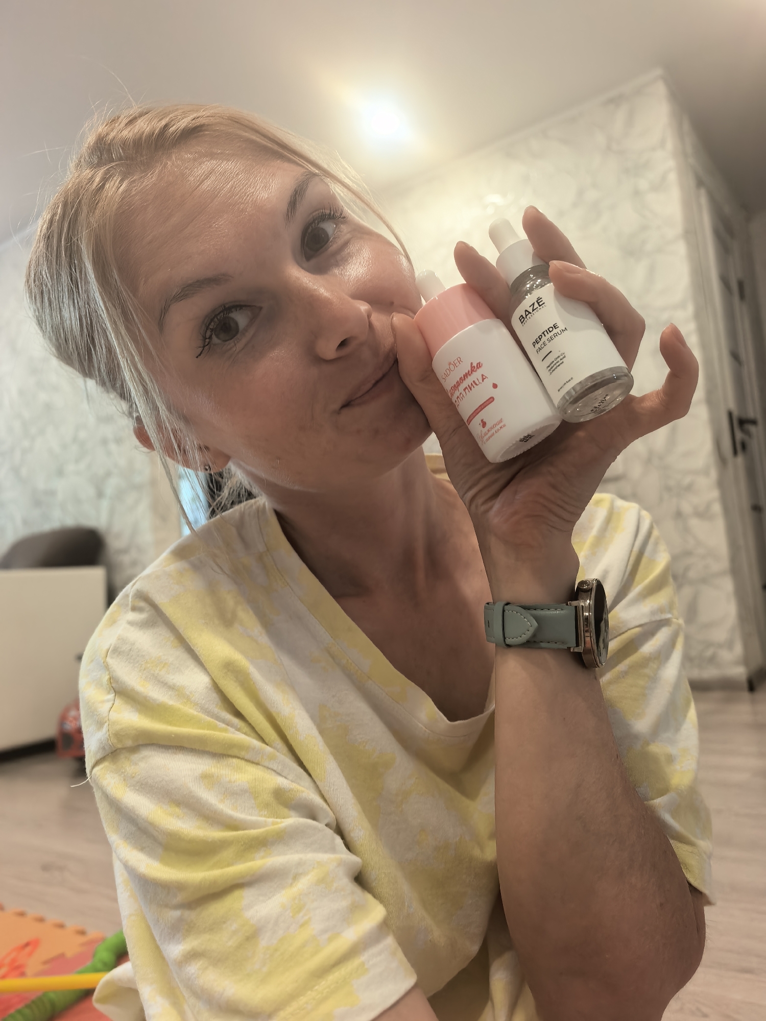 A woman with blonde hair pulled back is holding two cosmetic bottles in her right hand. The bottle on the left is pink and white, while the bottle on the right is white with a silver cap and contains "BAZE PEPTIDE FACE SERUM" written on the label. She is wearing a yellow and white t-shirt and a light blue watch on her left wrist. The photo was taken in Sterlitamak, Russia, inside an apartment, as evidenced by the background which includes a patterned wallpaper, furniture, and a children's play mat on the floor.
