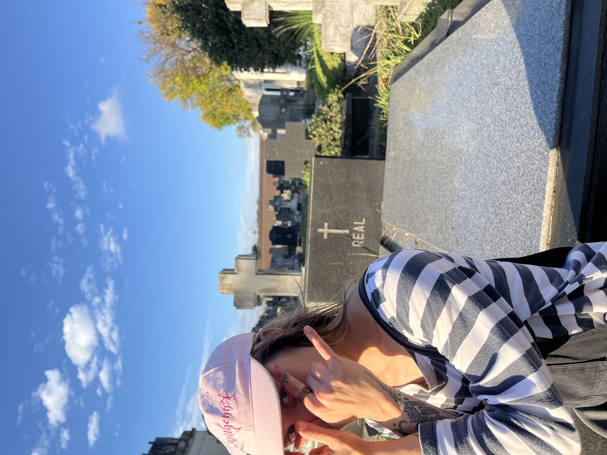 An image, originally oriented horizontally, captures a person posing in a cemetery under a clear blue sky. The subject, likely a woman, occupies the right side of the frame, shown from the chest up with her head slightly lowered. She wears a light pink baseball cap, featuring embroidered stylized text, possibly "Babygirl," on its front. Her hair is visible, and tattoos adorn her left forearm and hand. Her attire includes a long-sleeved shirt with distinct horizontal white and dark striped patterns, and dark clothing on her visible lower half.
Her right hand is raised, with the index and middle fingers extended upwards, partially obscuring her face around the eyes and nose. A metallic ring is visible on her right ring finger. Her left hand is also positioned near her face, further down.
The background depicts a cemetery, consistent with the provided location context of Olivos, Argentina. Numerous gravestones of varying sizes and designs, including several with crosses, are scattered across the landscape. A dark grey gravestone in the mid-ground features partially visible white text that reads "REAL" alongside a cross symbol. Green and yellow-leaved trees are visible on the left and in the distance. The sky is bright blue with scattered white cumulus clouds, indicating a sunny day. Strong sunlight casts visible shadows, suggesting mid-morning or afternoon. Due to her obscured face, the subject's emotions are not discernible.