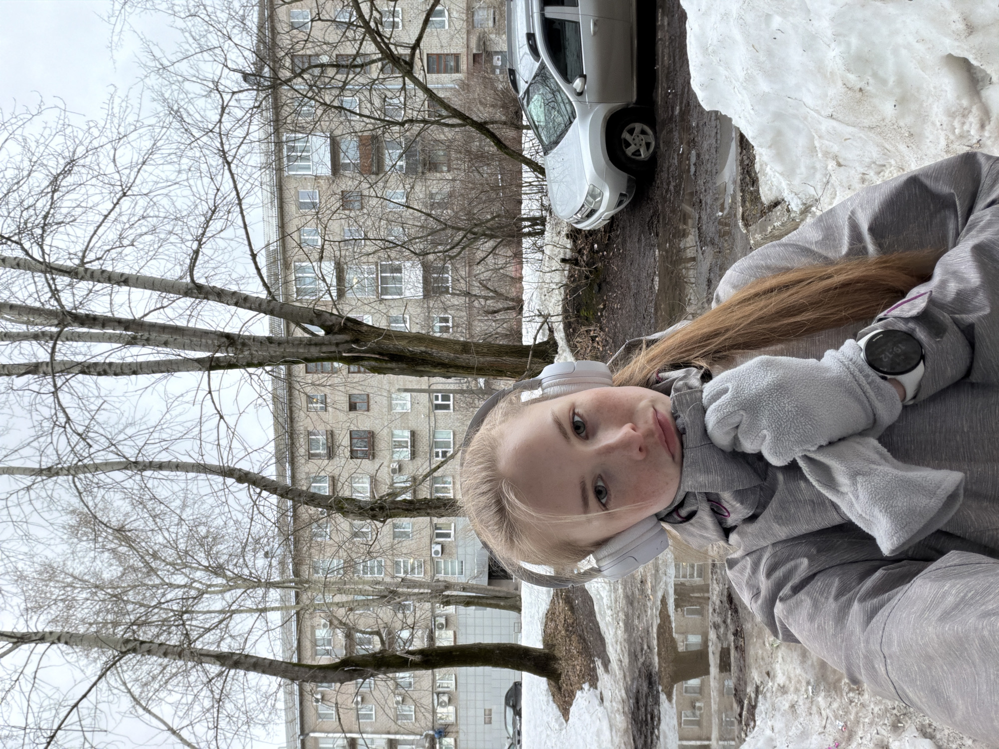 A selfie taken outdoors, likely in the city of Berezniki, Russia, during a season with melting snow. The subject is a young, light-skinned individual with blonde hair, wearing a grey jacket, gloves, and white headphones. A digital watch is visible on their wrist. In the background is a multi-story apartment building characteristic of Soviet-era architecture. A light-colored car is parked nearby. Bare trees suggest late winter or early spring. Melting snow is piled along the side of what appears to be a street or sidewalk.