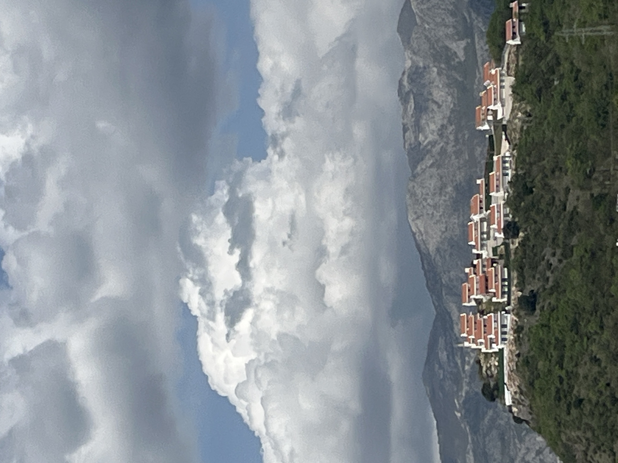 The image, taken in Blizikuće, Montenegro, presents a picturesque landscape featuring a row of residential buildings nestled into a verdant hillside. The scene is set under a partially cloudy daytime sky, with clear blue patches interspersed among large, soft white and gray clouds.
The primary subjects are the numerous multi-story buildings, characterized by their white facades and prominent red or terracotta-tiled roofs. These structures are arranged horizontally along a slope, suggesting a planned development, possibly a resort or residential complex. The hillside on which they are built is covered in lush green vegetation, including trees and shrubs, indicating a fertile environment.
In the background, a rugged, imposing gray mountain range rises majestically, forming a dramatic natural backdrop to the built environment. The texture of the mountains suggests rocky terrain.
There are no people or specific activities visible, emphasizing the architectural and landscape elements of the scene. The weather appears fair, characterized by bright daylight and the interplay of sun and cloud. The architecture, with its white walls and red roofs, is typical of Mediterranean and Adriatic coastal regions. No visible text is present in the image. The overall impression is one of a serene and integrated landscape where human habitation blends with striking natural beauty.
