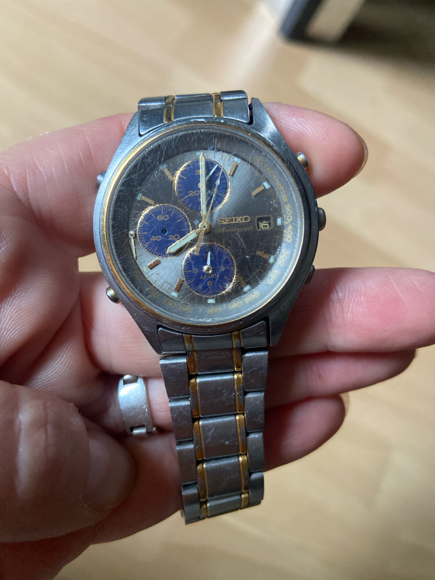 A wrist chronograph watch with a dark dial, gold accents, and three subdials is held by a person's hand. The watch has a metal band with gold links interspersed with silver links. The dial features blue subdials with white markings and gold hands. The bezel has tachymeter markings. The watch face shows some wear and scratches. The background is a blurred wooden surface. There are no identifiable landmarks or features that would suggest a location in Kassel, Germany.