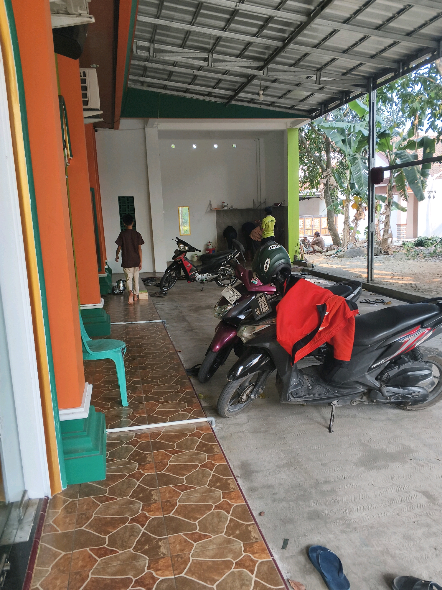 The image depicts an outdoor scene in Bandar Lampung, Indonesia, likely a residential or communal area with a covered walkway. In the foreground, a light blue plastic chair sits on a patterned tiled floor. To the left, an orange pillar with green and white accents is visible, part of a building's facade.
A young boy wearing a brown t-shirt and light-colored pants is walking away from the camera towards the right. Several motorbikes are parked in the background. One motorbike, predominantly black with red accents, has a red jacket draped over its handlebars and a green helmet with "INK" branding on the seat. Another motorbike, maroon in color, is parked beside it. A third motorbike, red and black, is also present.
In the middle ground, a person wearing a yellow shirt and helmet is visible near the motorbikes. Further in the background, under a metal-framed roof with a corrugated metal covering, more people are seated and interacting. Trees and foliage are visible beyond the covered area, suggesting an open-air environment. The ground surface varies from patterned tiles near the building to concrete or packed earth in the background. The overall impression is a casual, everyday setting in a warm climate.