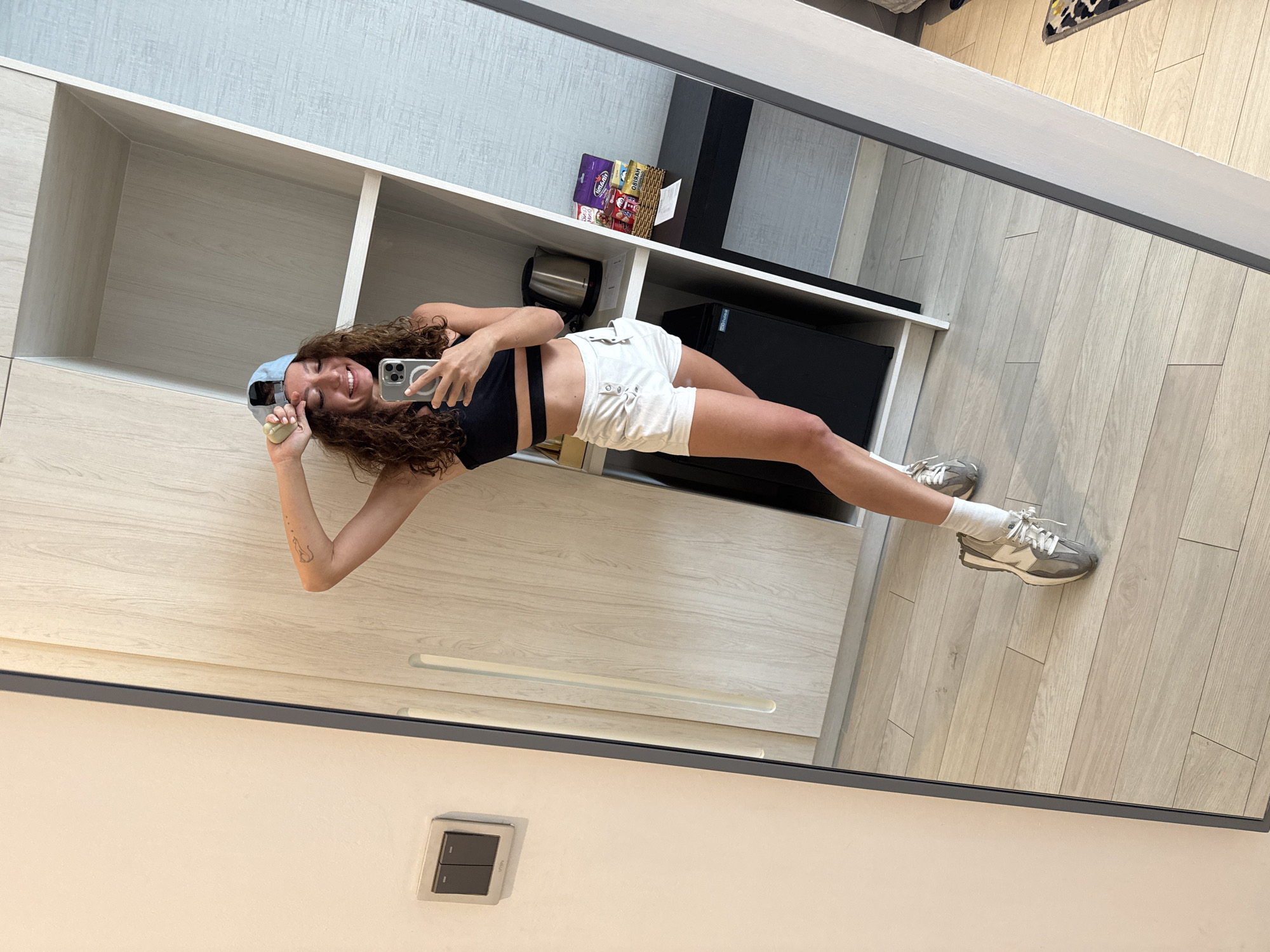 A young woman is taking a selfie in a mirror. She is wearing a black sports bra, white shorts, a blue baseball cap, white socks, and grey and white sneakers. She is standing in what appears to be a hotel room. The room has light wood-colored floors, white walls, and a built-in shelving unit. The shelving unit contains a small refrigerator, a coffee maker, and some snacks. A light switch is visible on the wall below the mirror. The room does not provide enough detail to identify the location in the city of Đà Nẵng, Vietnam.
