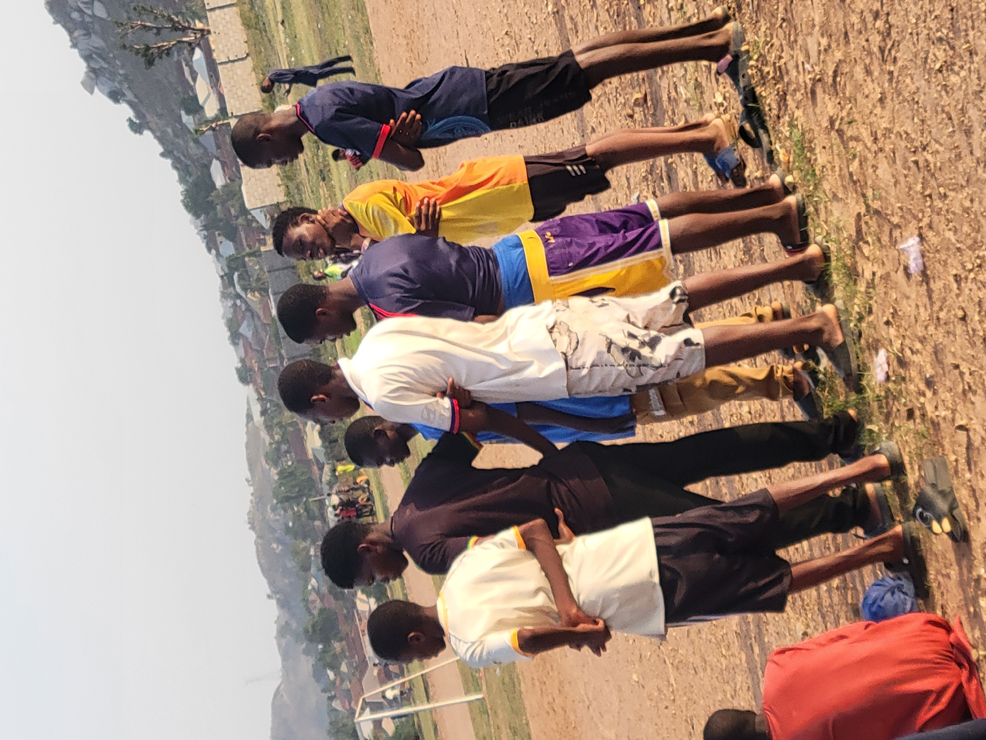 A row of seven Black African men, wearing various casual athletic clothing including shorts, appear to be performing some form of a bow or prayer on a dirt field. Behind the row is a slightly blurred view of a cityscape at the base of a rock outcropping. There is a red bag next to the last person in the row. The overall area appears to be on the outskirts of a developed area in Ibo, Nigeria.