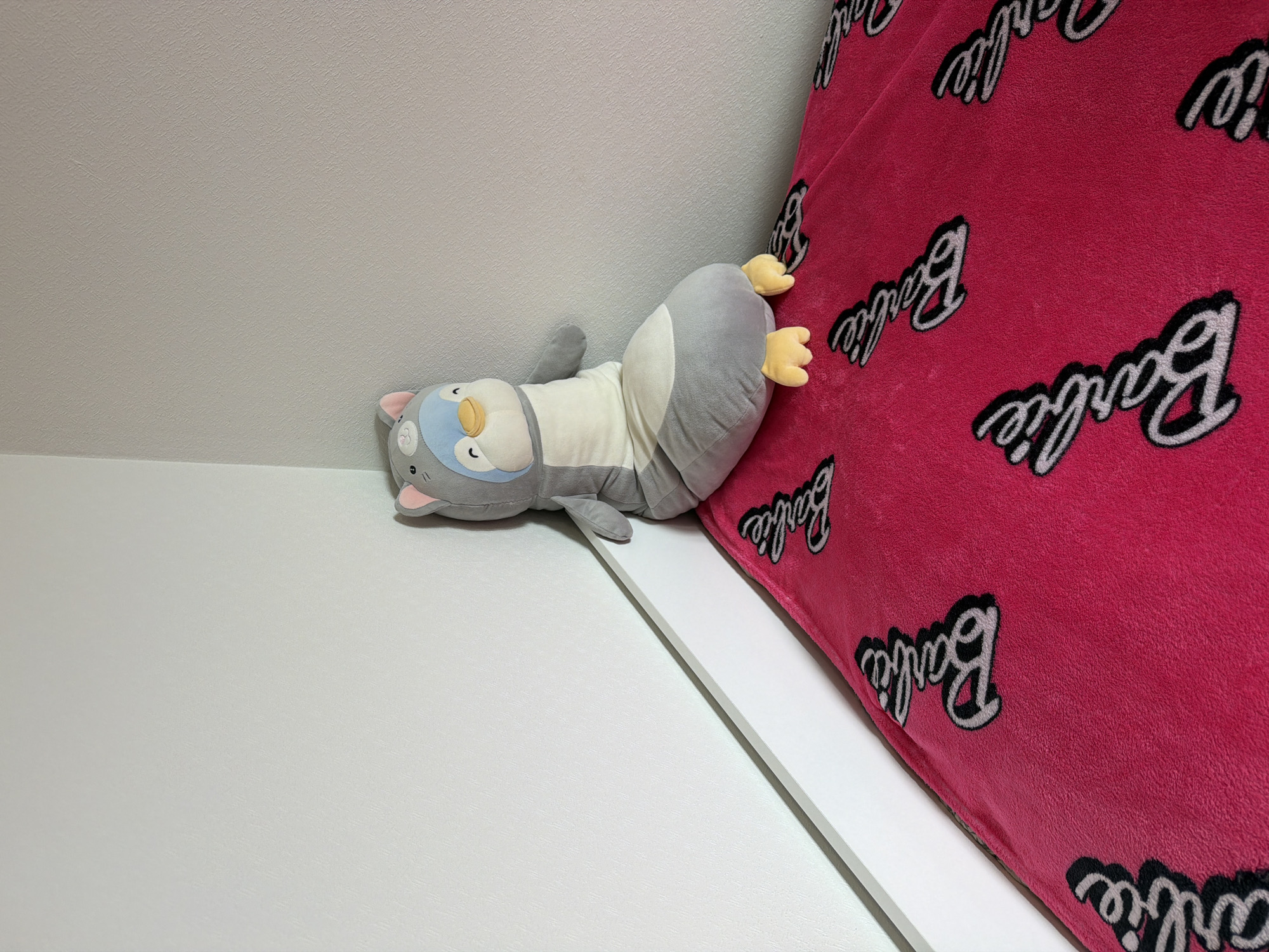 The image displays an indoor scene featuring a plush toy and a blanket. A grey and white plush toy, resembling a penguin or a small animal with a light blue face, yellow beak, and yellow webbed feet, lies on its back. It has small pink inner ears. The toy is positioned on a flat, white surface, possibly a bed frame or shelf, and rests against a light-colored, textured wall. Adjacent to the plush toy, a vibrant pink blanket is draped over the white surface, leaning against the wall. The blanket is patterned with the word "Barbie" repeated several times in a stylized black and white script font. The wall exhibits a subtle, diagonally-oriented textured pattern, while the blanket appears soft and fleecy. The scene is well-lit, likely by indoor artificial light. There are no discernible activities taking place, and no emotions are depicted by the inanimate objects. No specific location cues for Moscow, Russia, are visible within the image itself.