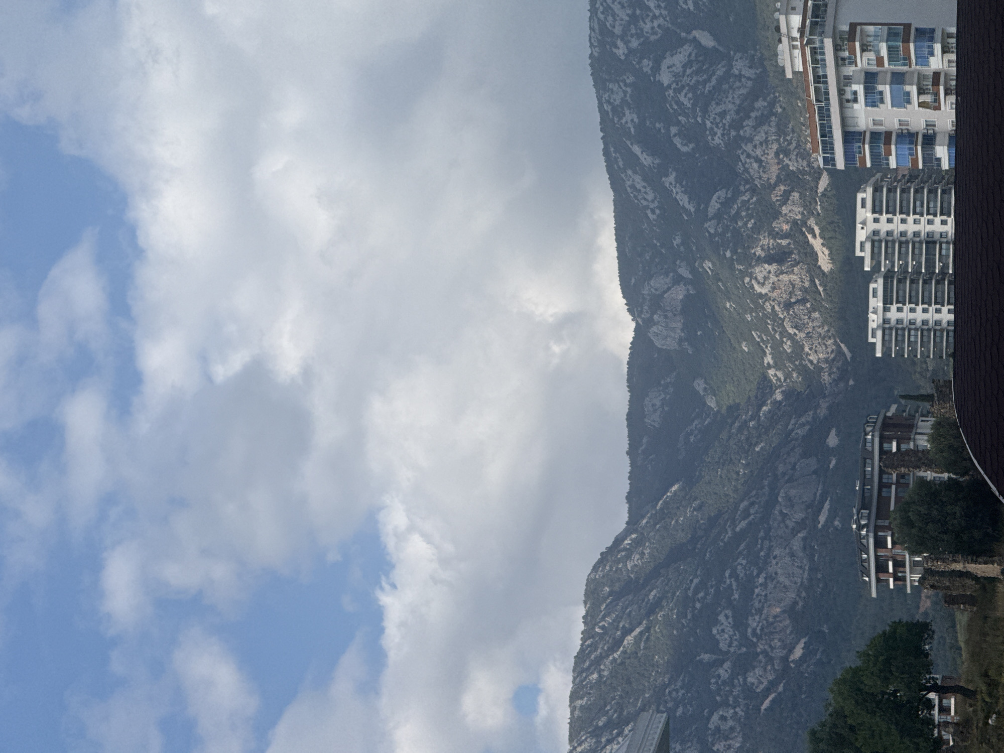 The image depicts a mountain range bordering a city. The mountain is tall and rugged, with exposed rock and sparse vegetation. Buildings line the base of the mountain, including high-rise apartments with multiple stories and windows. The sky is partially cloudy with patches of blue. The location is unknown.