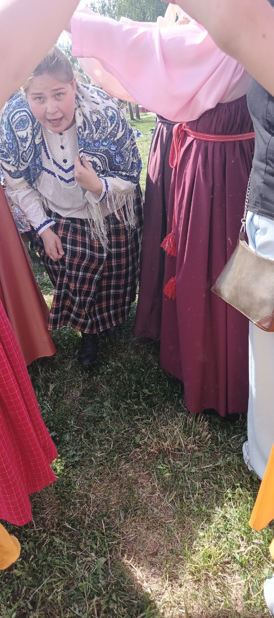 In Кемуль, Russia, a group of people wearing traditional clothing stand on a grass lawn. A woman in the center wears a white shirt with blue trim, a blue and white patterned shawl, and a plaid skirt. A red skirt, a maroon skirt with a red rope belt, and a light blue skirt are visible around her.