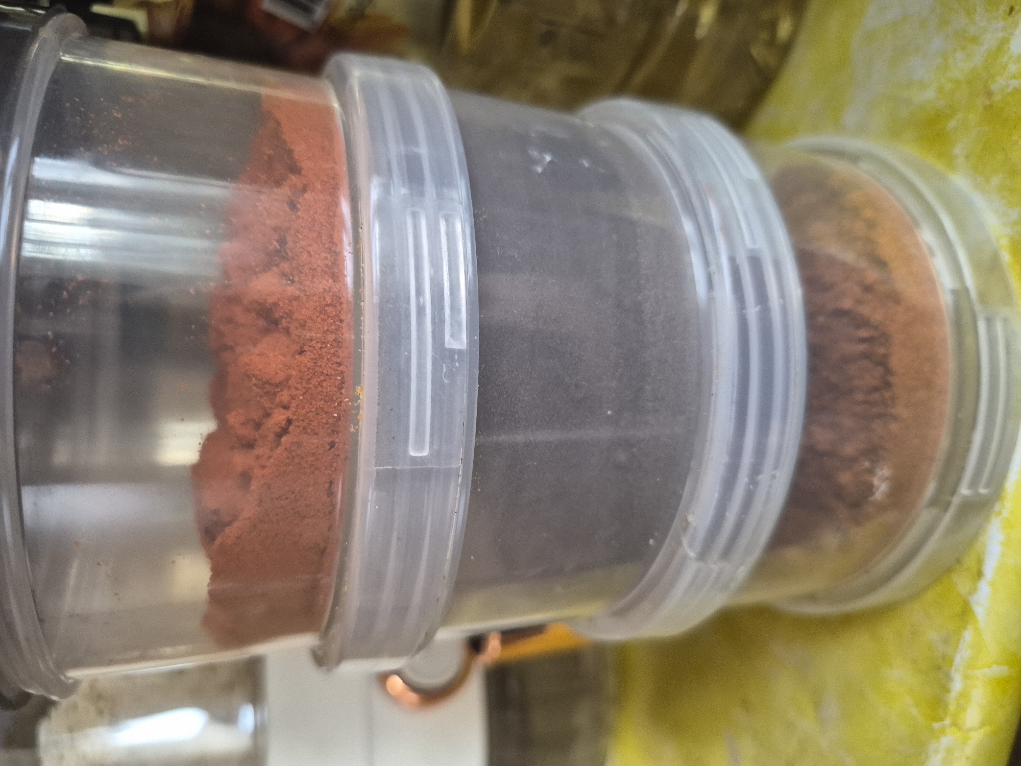 The image displays a close-up, horizontal view of three interconnected clear plastic cylindrical containers, likely used for storing spices. Each container holds a different finely ground powder.
The leftmost container is filled with a vivid reddish-orange powder, reminiscent of paprika or chili. The middle container contains a dark grey to black powder, which could be black pepper, a dark herb, or another ground spice. The rightmost container holds a lighter brown or orange powder, possibly cumin, coriander, or a similar spice. The clear plastic material of the containers allows the colorful contents to be easily seen, and the containers appear to screw into each other with ribbed plastic rings, forming a stackable, modular system.
The background is softly blurred, showing hues of yellow and lighter neutral tones, suggesting an indoor setting such as a kitchen, pantry, or a retail display in Yerevan, Armenia. A faint, curved metallic object, possibly made of copper, is visible beneath the central container. No specific activity is taking place; the image is a static portrayal of stored ingredients. There is no visible text or branding on any of the containers or in the surrounding environment. The lighting is consistent with indoor ambient light.