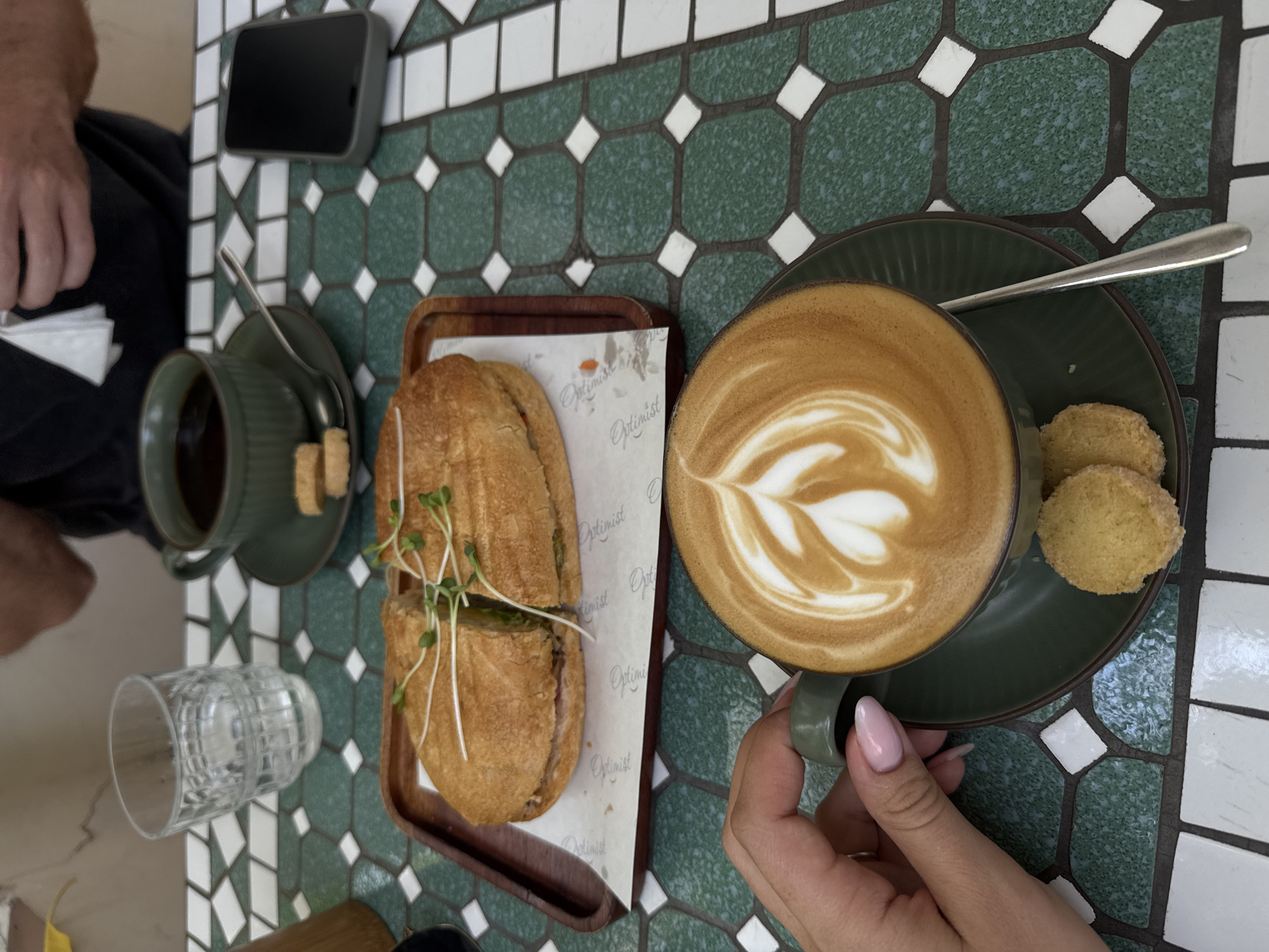 The image shows an overhead shot of a table setting in an outdoor cafe or restaurant, likely in Unknown, Unknown. The table has a green and white tiled surface. A person is sitting at the table, evidenced by a partial view of an arm, hand with pink nail polish holding a green cup filled with a latte featuring latte art, and part of another person's arm and leg is seen on the left side.
On the table are the following items: a dark-colored smartphone lying flat, a green teacup filled with dark liquid and a spoon on its saucer, a clear glass of water with remnants of water drops, a wooden tray holding a sandwich cut in half and garnished with sprouts, and a small green plate with the latte cup and two cookies. The sandwich is placed on a white piece of paper with the word "Optimist" printed on it.