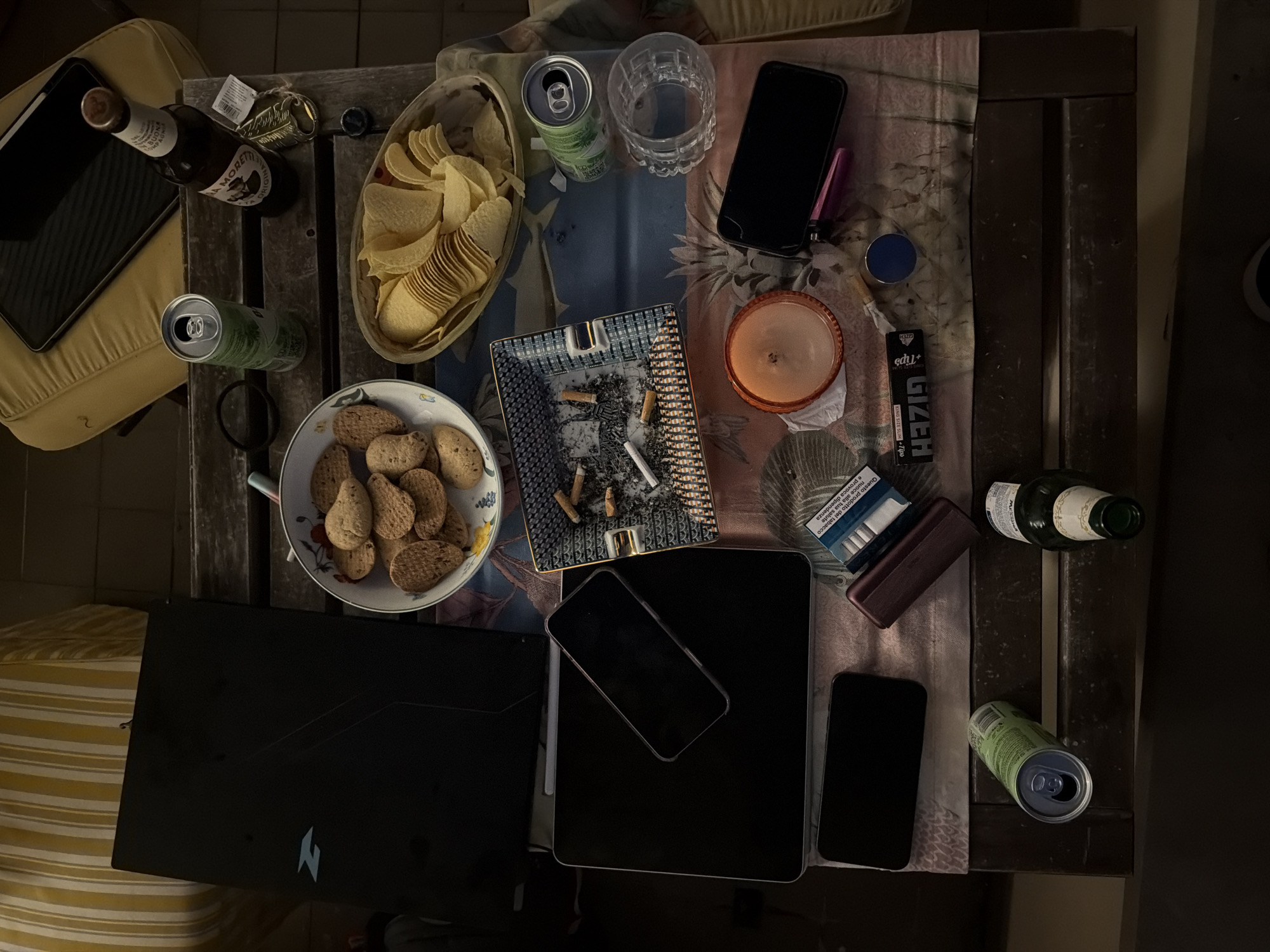 The image presents an overhead view of a wooden table, possibly a patio or balcony setting, partially covered by a patterned tablecloth. The dim lighting suggests it is evening or night. The scene depicts a casual, relaxed atmosphere, possibly involving a social gathering or an individual unwinding.
The table is cluttered with various items. Food includes a bowl of potato chips and a white bowl filled with small, light brown biscuits. Beverages consist of an empty Birra Moretti beer bottle, displaying "BIRRA MORETTI ORIGINAL BUONA COMPAGNIA" on its label, two open green cans, an empty clear glass, and a dark glass bottle.
Smoking-related items are prominent: a patterned ashtray filled with cigarette butts and ash, a blue and white cigarette packet with the Italian health warning "Questo prodotto del tabacco nuoce alla tua salute e provoca dipendenza" (This tobacco product harms your health and causes addiction), a brown cigarette case, and a packet of "HAZID eDU+ KING SIZE SLIM+Tips" rolling papers.
Electronics include a black laptop (with a visible Acer Predator logo), a black tablet, and three black smartphones, one of which appears to have a cracked screen. Other items on the table are a small, orange candle in a glass holder, a blue circular tin, a pink object, and a black hair tie. A light yellow cushion is partially visible to the left. The arrangement suggests a period of relaxation or informal activity, consistent with a home setting in a city like Milan, Italy.