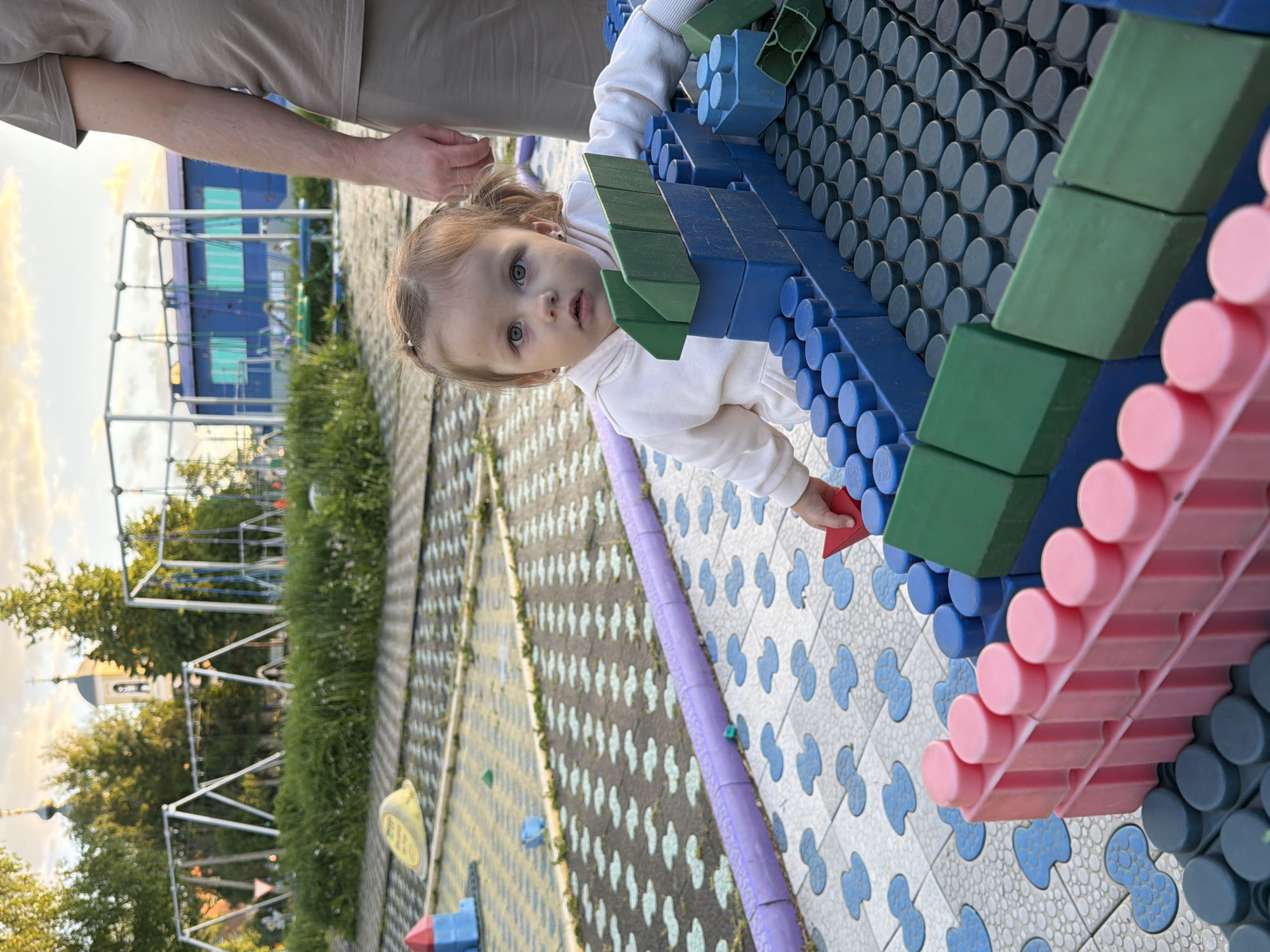The image shows a young child with light hair and a white sweater, looking at the camera while playing with large, colorful building blocks made of blue, green, and pink plastic in a playground. An adult's arm and hand are partially visible above the child. The ground is covered with patterned tiles in gray and blue. In the background, there are trees, a playground structure, and a building. A church steeple is visible among the trees. The photo was likely taken in Sin'kovo, Russia.