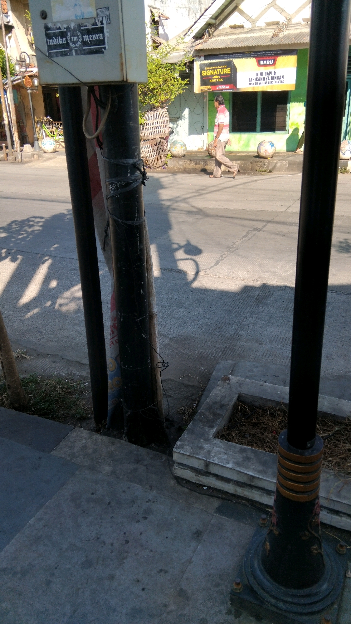 The image captures a street scene in Batang, Indonesia, likely in the morning due to the long shadows cast by the objects and buildings. In the foreground, a black lamppost with decorative gold rings stands on a concrete base, anchoring the right side of the frame. To the left, another dark pole is visible, partially obscuring a weathered utility box with stickers on its upper section, one of which reads "tadika tm mesra". Wires are visibly taped to the pole.
In the midground, a man walks away from the camera, wearing a light-colored collared shirt with pink stripes and beige trousers. He is on a paved sidewalk that runs parallel to the street. Behind him, a green building with a sign above it advertising "BARU" (New) and "SIGNATURE KRETEK" is prominent. Next to the building are large, round woven baskets and some decorative spherical objects on the ground. A bicycle with a green frame is parked to the left of these elements. The street itself is paved and shows signs of wear. The overall environment suggests an urban or semi-urban setting with commercial activity.