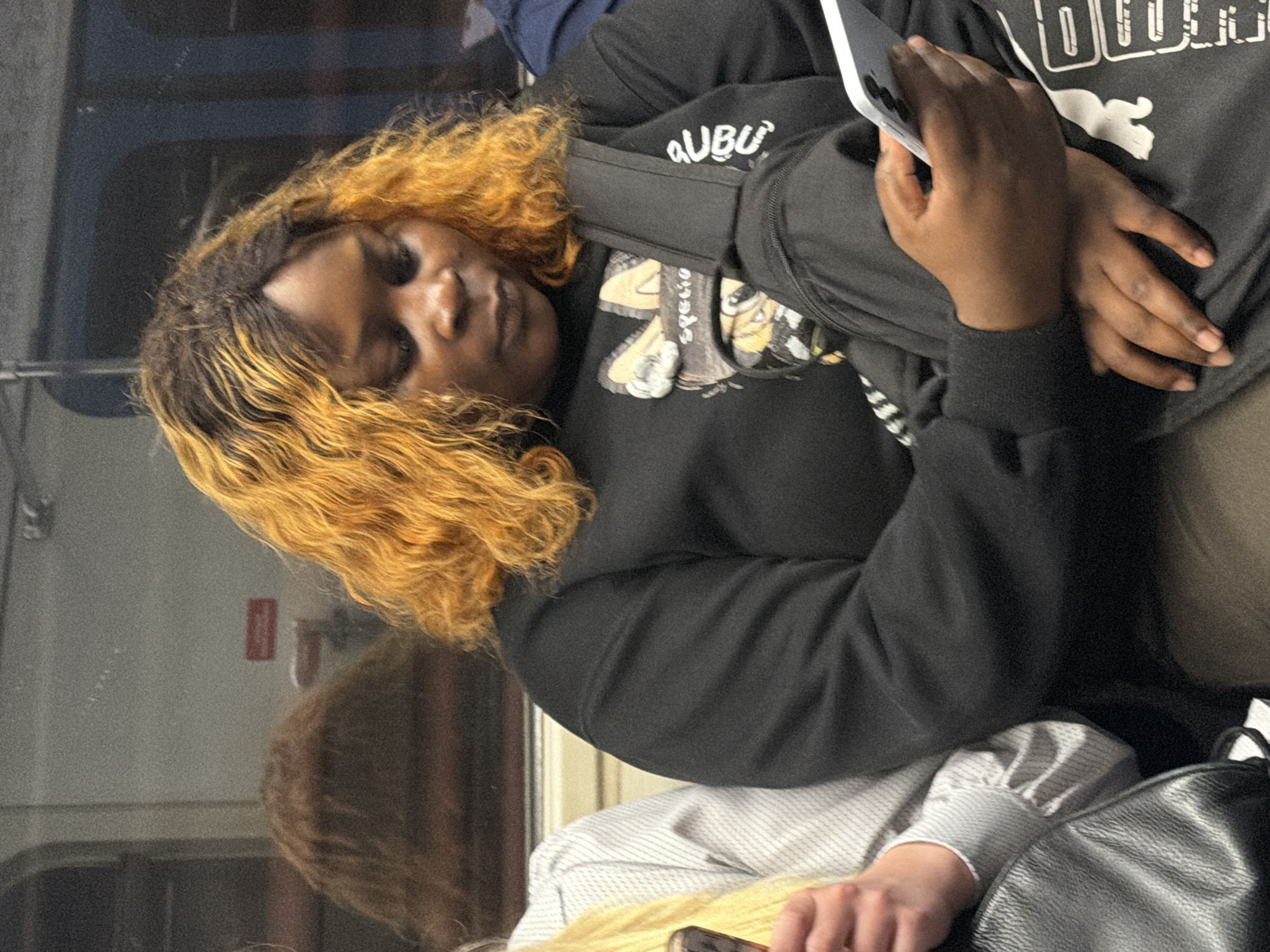 A person with orange-tinted hair is seated indoors, holding a smartphone in their right hand. They are wearing a black sweatshirt with a graphic on the front. Another person with blonde hair is partially visible to the left, also seated. The background appears to be a window with reflections, suggesting a form of public transportation or a room with a large window. The lighting is dim.