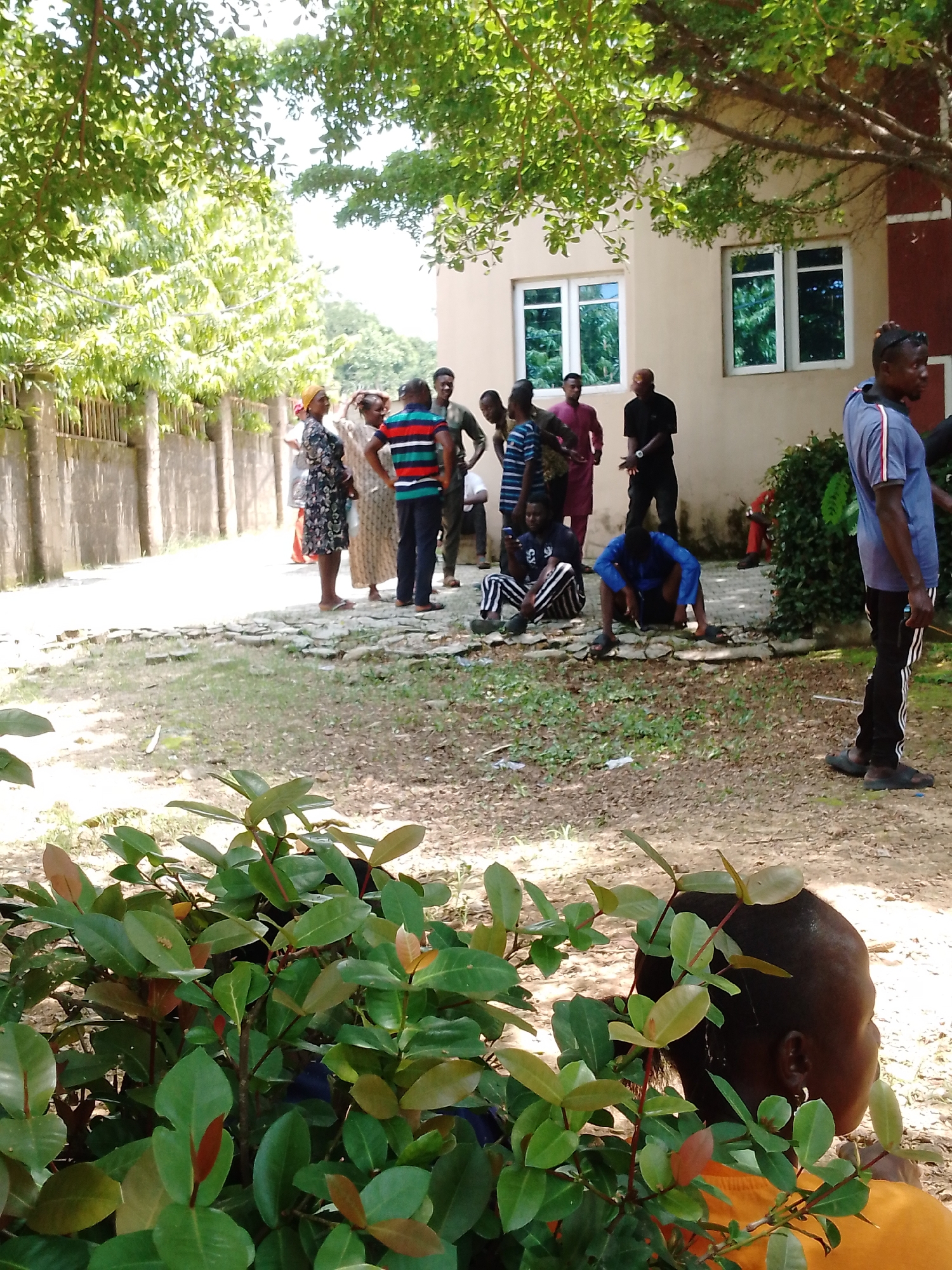 The image depicts an outdoor gathering of approximately ten people, primarily adults, in what appears to be a compound or yard area. In the foreground, lush green foliage obscures the lower portion of the scene. Behind the plants, several individuals are visible standing and sitting.
Towards the left, a group of people are gathered near a textured wall and a fence. A woman in a patterned dress and headscarf stands out, next to others in casual attire. Further right, a building with light-colored walls and two sets of windows is visible. A man in a blue shirt with red and white stripes is standing and looking towards the right. Other individuals are seen standing or interacting near the building entrance.
In the mid-ground, two men are seated on the ground amidst scattered stones and dirt. One is wearing a blue top and dark pants, while the other is in black and white striped pants and a dark top. Another individual in a red shirt is seated further back, partially obscured by a bush.
The environment suggests a warm climate, with ample sunlight filtering through the trees. The scene appears to be a public or residential area, possibly a community gathering or a waiting area. The presence of the building and the outdoor setting implies a location in Akwanga, Nigeria, likely a non-commercial or communal space. The interactions among the individuals are not clearly defined, but they appear to be in a relaxed, informal setting.