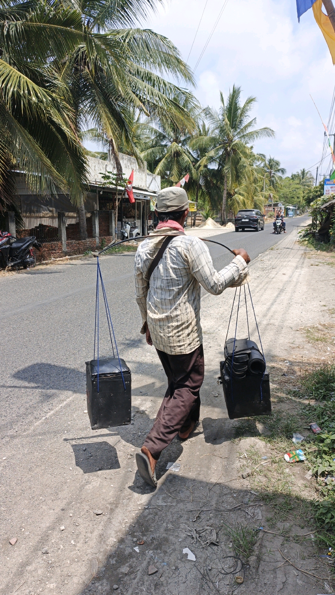 A man walks along a dirt path beside a paved road under palm trees in Desa Karangjaladri, Indonesia. The man, wearing a light-colored checkered shirt, dark pants, a tan hat, and a red cloth around his neck, carries two black, box-like containers suspended from a long pole balanced on his shoulders. Blue ropes attach the containers to the pole. The left container is closer to the man and casts a square shadow. The right container is further away and has rolled items, possibly mats, inside. A dark SUV is visible driving on the road in the background, along with a motorcycle with two riders. Buildings with red and white flags are on the left side of the road, and vegetation lines the right side. The sky is partly cloudy.