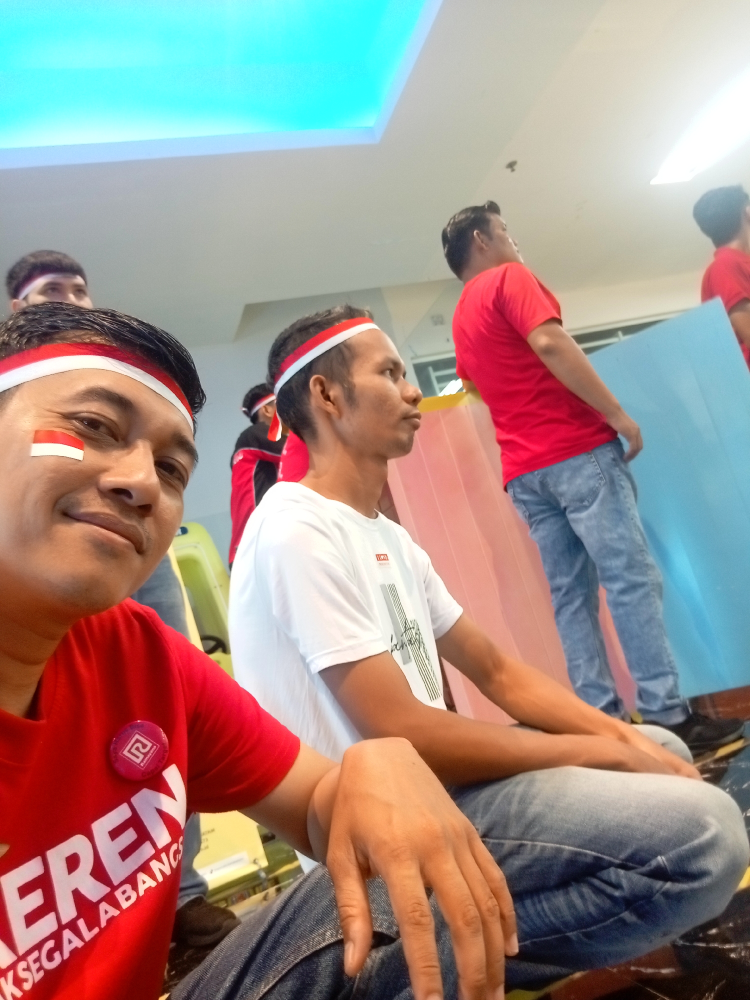 In Batam, Indonesia, a group of men in red t-shirts and white headbands with red stripes are seen. One man in the foreground is smiling and has the Indonesian flag painted on his cheek. He is wearing a red t-shirt with white text and a circular pin. Another man in the mid-ground is wearing a white t-shirt and blue jeans, facing right. In the background, several other men in red shirts are visible, some holding a light blue object. The environment appears to be indoors with a bright, aqua-colored ceiling light.