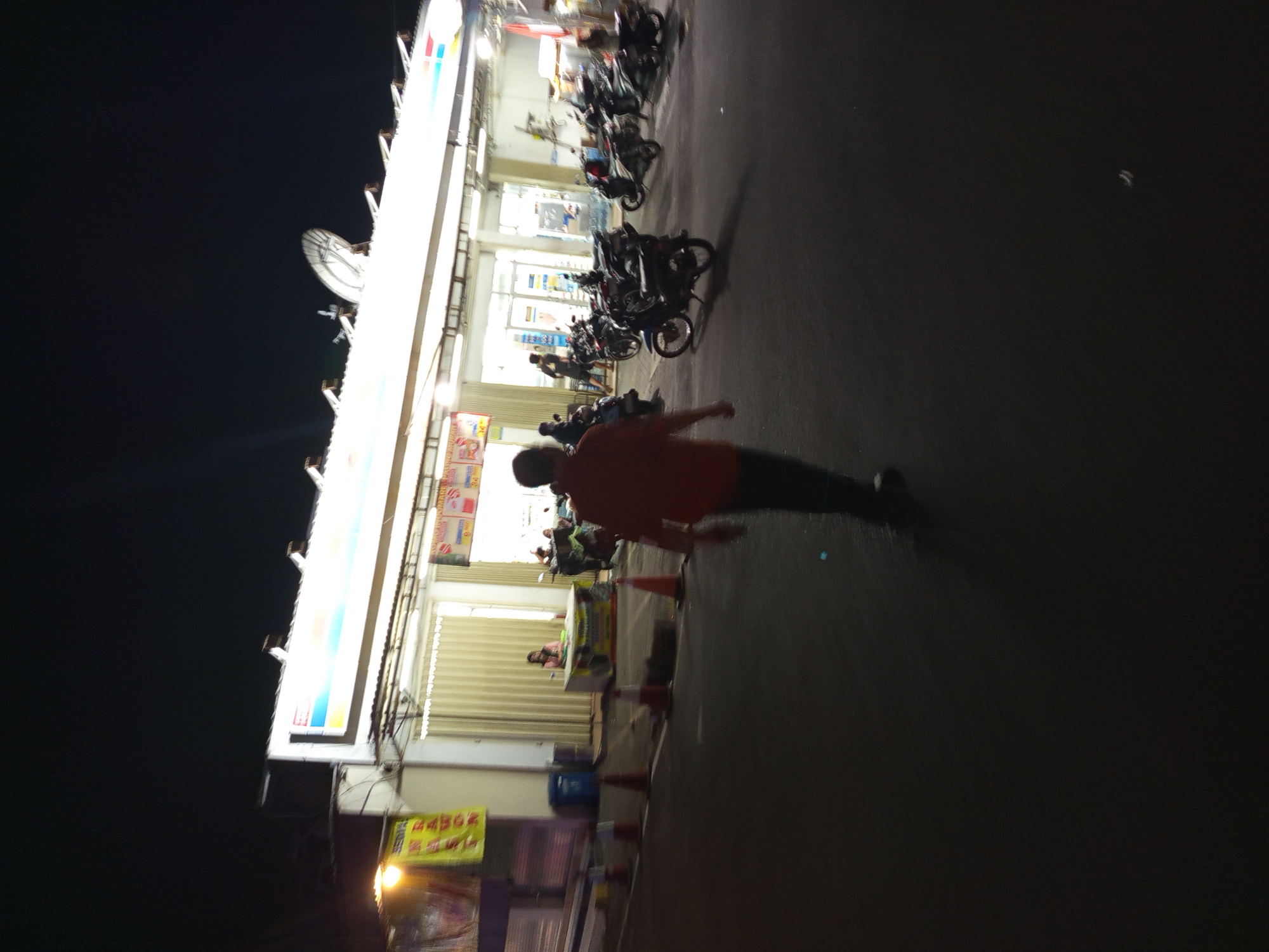The image depicts a street scene at night in Gemurung, Indonesia, likely in front of a convenience store. A man in a red shirt is walking away from the camera towards a brightly lit storefront. Several motorcycles are parked in front of the store, and a woman is visible behind a counter inside. The street is paved, and traffic cones are placed along the side. The sky is dark, indicating nighttime.
