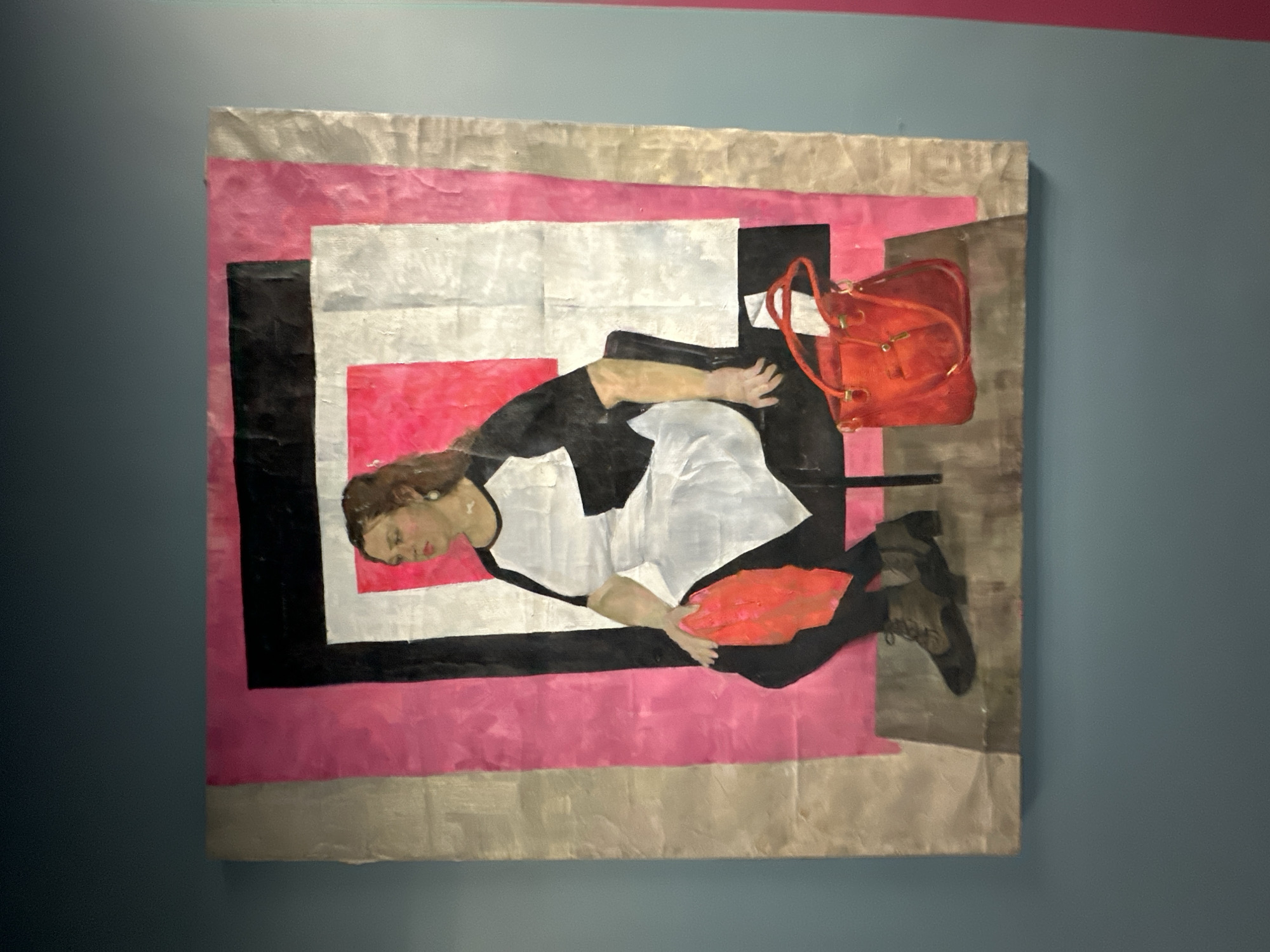 A painting hangs on a blue wall with a pink stripe near the top. The painting features a reclining figure against a geometric background. The figure, a woman, is in a black and white dress with black leggings and boots. Her head is turned to the left and eyes closed. Her left hand is resting on a red area of the painting. A red purse with straps is positioned near her torso. The geometric background is composed of pink, white, gray, and black rectangles. The location is Saint Petersburg, Russia.