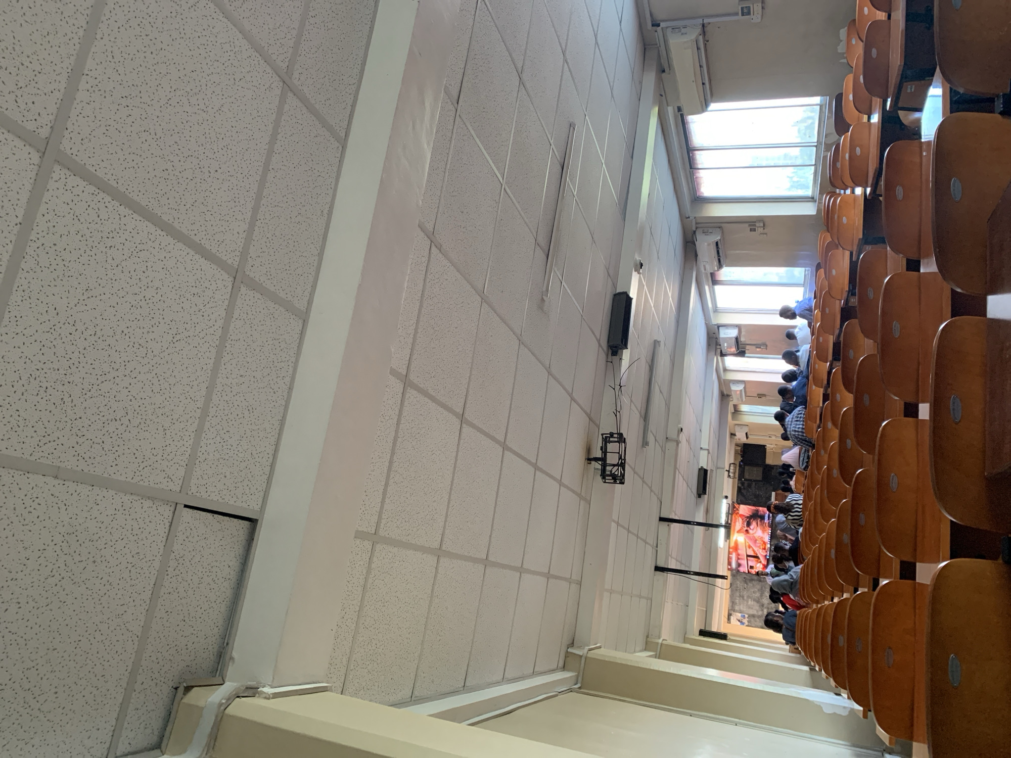 The image captures the interior of a large hall, likely a lecture room or auditorium, viewed from a slightly elevated position and rotated clockwise, causing the ceiling to appear on the left.
The left side of the frame is dominated by a suspended acoustic tile ceiling, featuring a grid pattern of light gray panels. White structural beams or columns run vertically and horizontally across this section. Further right, along what would be the upper wall, are white walls with mounted black speakers and metal brackets.
On the right side of the image, rows of light brown wooden chairs with slightly darker seat and back pads are arranged in a tiered auditorium style. Numerous people are seated in these chairs, facing towards the far end of the room. Most individuals are seen from behind or in profile, dressed in casual attire such as shirts, some with plaid patterns.
At the far end of the room, several large rectangular windows provide natural light, indicating it is daytime. Two wall-mounted white split-type air conditioning units are visible beneath these windows. The people are focused on a large screen or television display at the front of the room, which is showing a vibrant, colorful image that appears to depict figures, possibly from a cultural or religious context.
The scene suggests an ongoing event such as a lecture, presentation, or communal gathering where an audience is observing content displayed on a screen. The overall ambiance is one of calm attention. No discernible text is visible within the image. Given the provided context, this setting is located in Dar es-Salaam, Tanzania.