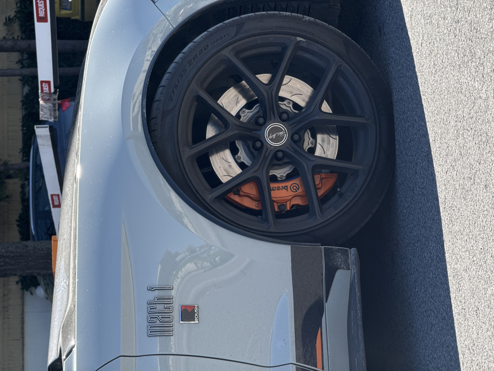 A close-up view of the front driver-side wheel and body panel of a light-colored Mach 1 Roush Mustang. The wheel is black with a multi-spoke design, revealing Brembo orange brake calipers. The tire is marked "275/35 ZR20 (102Y)". The Mach 1 logo and Roush badge are visible on the body panel. The image appears to be taken outdoors on a paved surface. There is no evidence to indicate that the location is in Plovdiv, Bulgaria.