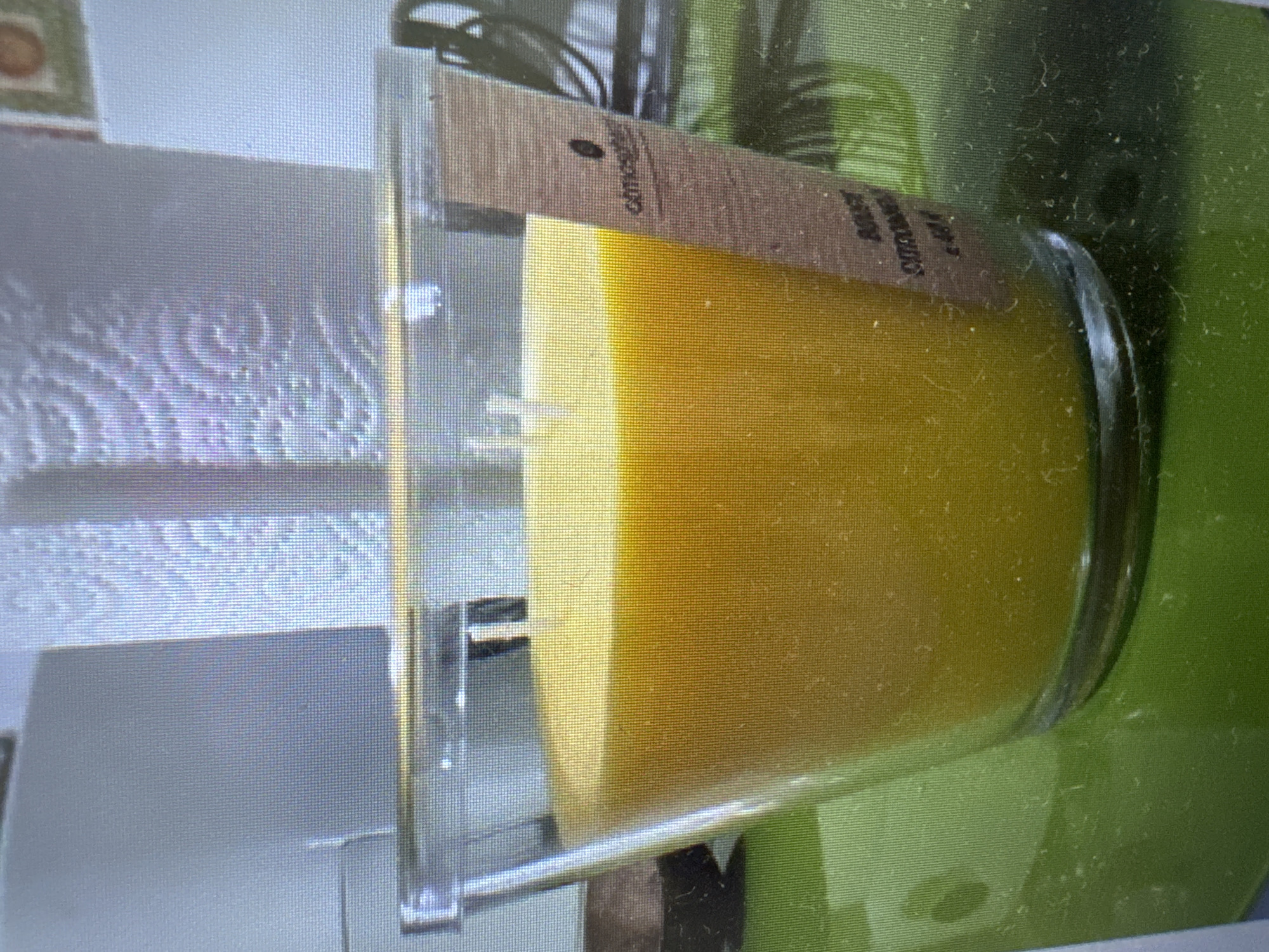 The image shows a close-up of a yellow candle in a clear glass container, possibly a large jar candle. The candle has a visible wick. A small, rectangular label is attached to the side of the glass, obscuring part of the candle's surface. The label is light brown with black text. The text "office" is visible on the left side of the label, and other text, appearing to be "ROSE CITRONELLE", is visible on the right. The candle and its container are placed on a reflective surface, possibly a table. The background is out of focus and appears to be a mix of green and gray tones, suggesting an indoor setting, possibly a room with plants or a green surface. The lighting suggests it is daytime, but no specific time or weather conditions can be determined. There are no people visible in the image. The overall impression is that of a product photograph or a detail shot of a home decor item.
