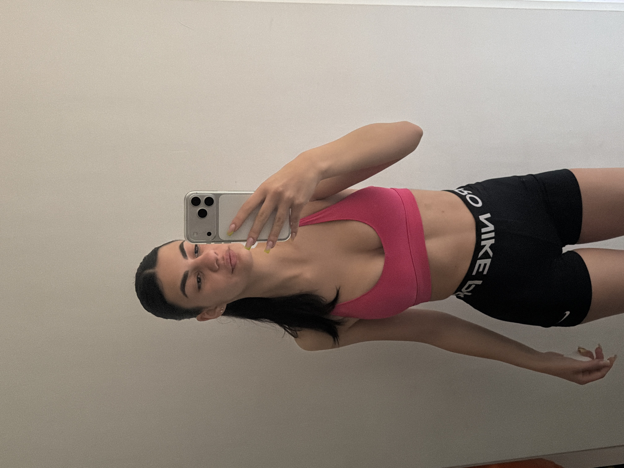 A young woman is visible in a medium shot, positioned against a neutral-toned wall. She is facing the camera and holding a silver smartphone up with her right hand, presumably to take a selfie. The woman is wearing a bright pink sports bra and black athletic shorts with the word "NIKE" in white lettering around the waistband. Her dark hair is pulled back. She has fair skin and is looking directly at the camera. The setting seems to be indoors, likely in a room with a minimalist design. The location is unknown and no landmarks indicate a presence in Gabrovo, Bulgaria.