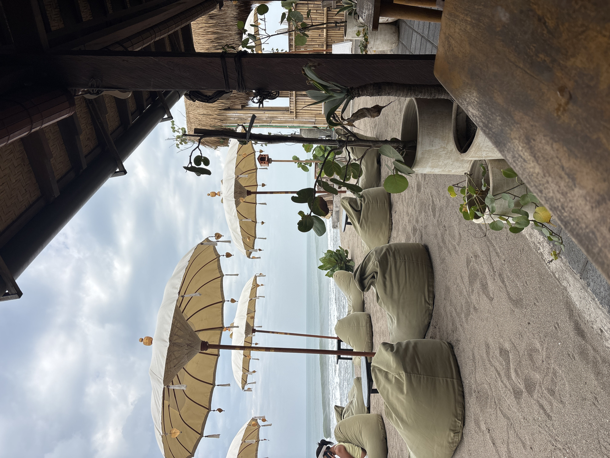 A beachside lounge area in North Kuta, Indonesia features several large, pale yellow beach umbrellas providing shade over scattered olive-green beanbag chairs on a sandy shore. The ocean is visible in the background, with moderate waves under a cloudy sky. To the right, a thatched-roof structure with wooden pillars and bamboo walls suggests a cafe or bar. Potted plants, including one with broad green leaves and another with spiky foliage, are placed on the paved area near the structure. A wooden table occupies the foreground, partially obscuring a clear view of the beanbag seating. One person is visible sitting on a beanbag near the ocean. The scene depicts a relaxed outdoor seating arrangement at a coastal establishment.