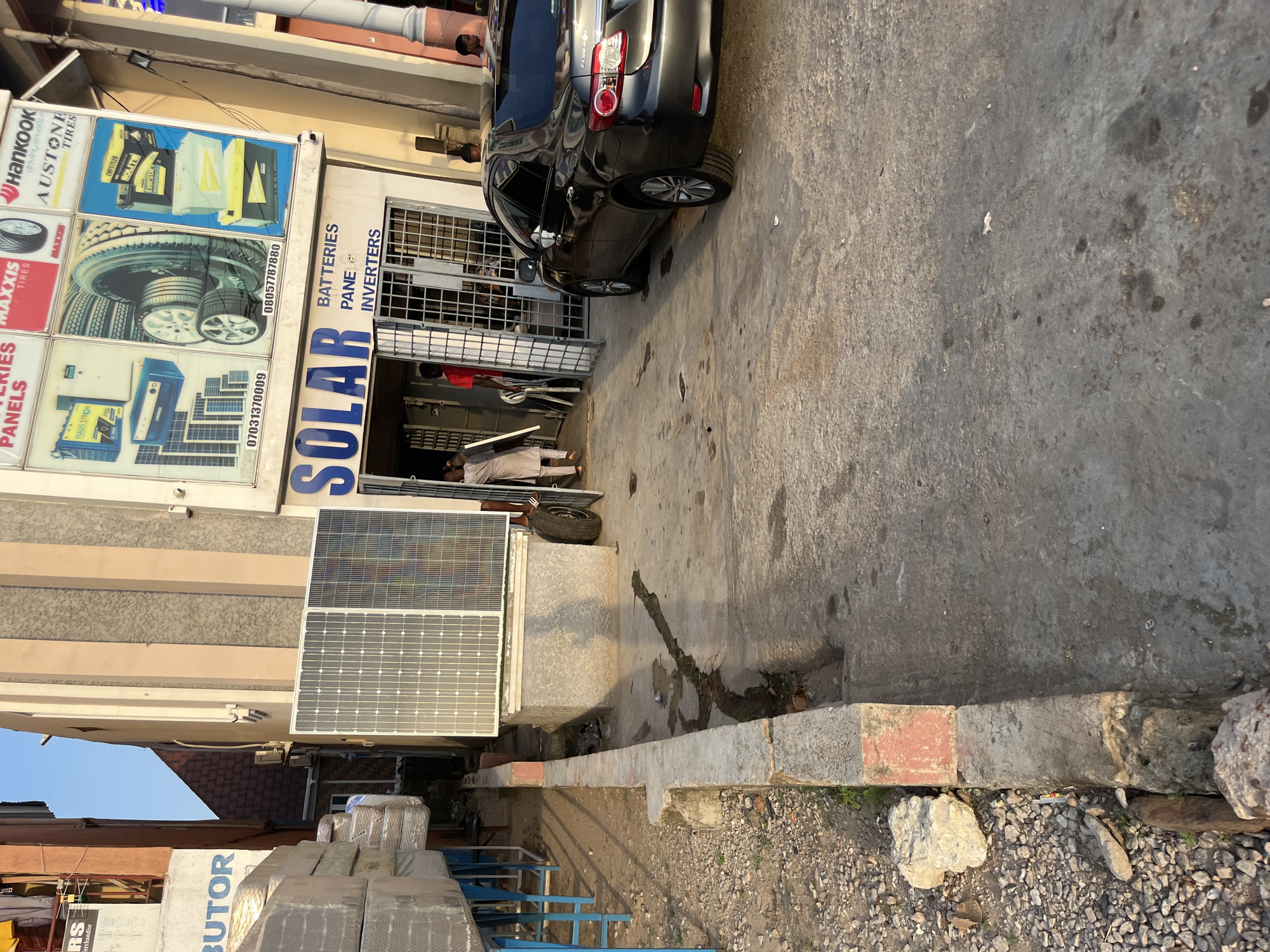 In an outdoor urban setting, a shop displays "SOLAR BATTERIES PANE INVERTERS" above its entrance. The storefront features a metal gate and two solar panels are mounted on the wall above and to the left of the entrance. Adjacent to the shop, a dark-colored sedan is parked. To the left of the shop, stacks of mattresses are displayed on blue stands. The ground is paved with concrete, and a drainage channel runs along the side. Several individuals are visible, including one standing in the doorway and others inside the shop. The sky above is clear and blue.