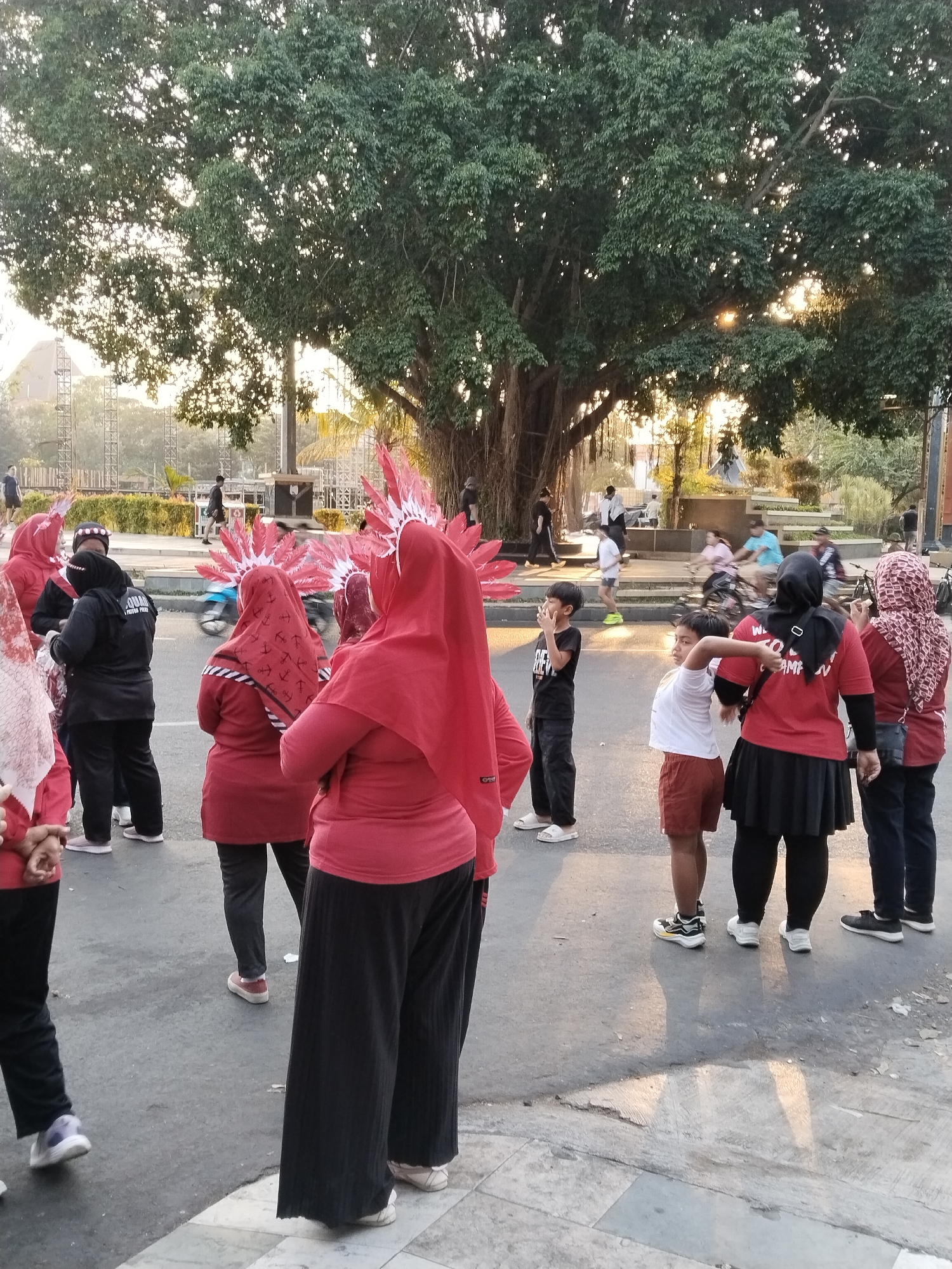 The image depicts an outdoor scene, likely in a public space in Kecamatan Pati, Indonesia, during daylight hours, with the sun casting long shadows. A large, mature tree with extensive roots and a dense canopy dominates the background, its branches allowing sunlight to filter through. In the foreground and midground, a group of individuals are gathered on a paved street.
Several people are dressed in matching red attire, including headscarves. Some are wearing elaborate red feather headdresses. One person in the foreground wears a black jacket over a red shirt and black pants, with a logo visible on the jacket. Another individual, a child in a black t-shirt, stands facing away from the camera. A younger child in a white t-shirt and red shorts is captured in profile. Other individuals in red tops and black pants are also visible, some appearing to be engaged in an activity or preparing for one.
In the background, a street with a few vehicles, including a motorcycle, is present. Further back, there are structures that appear to be part of a stage or event setup, with scaffolding and large screens. In the distance, other people are scattered throughout a park-like area with greenery and some architectural elements. The overall impression is of a community gathering or event taking place outdoors.