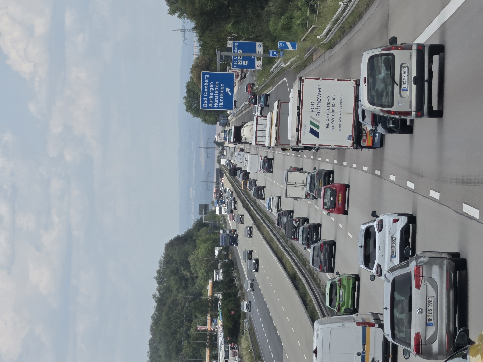 The image depicts a heavily congested highway with multiple lanes of traffic in Germany, specifically on a section of the A3 autobahn heading towards Bad Camberg-Kernstadt. Numerous cars, trucks, and a few vans are stopped or moving very slowly. The sky is overcast with scattered clouds.
In the foreground, a silver Volkswagen Passat station wagon with license plate "ME OS 1981" is prominently visible. To its right, a white Ford Puma ST with license plate "AW JM 607" is also in frame. Behind these cars, other vehicles including a silver van with license plate "LM MX 500" are present.
Further down the highway, a large white truck with the branding "von schaewen" is stopped in traffic. Other trucks, some with trailers and others articulated, are also a significant part of the scene. The highway infrastructure includes guardrails and a concrete median.
On the right side of the highway, a blue highway sign indicates directions to "Bad Camberg", "Aarbergen", "Hünfelden", and "Hünfelden", with an arrow pointing to the right. Below this, smaller signs include an "M" for a McDonald's, a "U70" sign, and a "100 km/h" speed limit sign.
On the left side of the highway, a gas station with multiple pumps and convenience store is visible in the distance, along with a line of parked trucks. The surrounding landscape features a dense forest on a hill. Overhead, power lines and pylons are visible against the sky. The overall scene captures a typical traffic jam on a German autobahn, likely during peak hours or due to an incident.