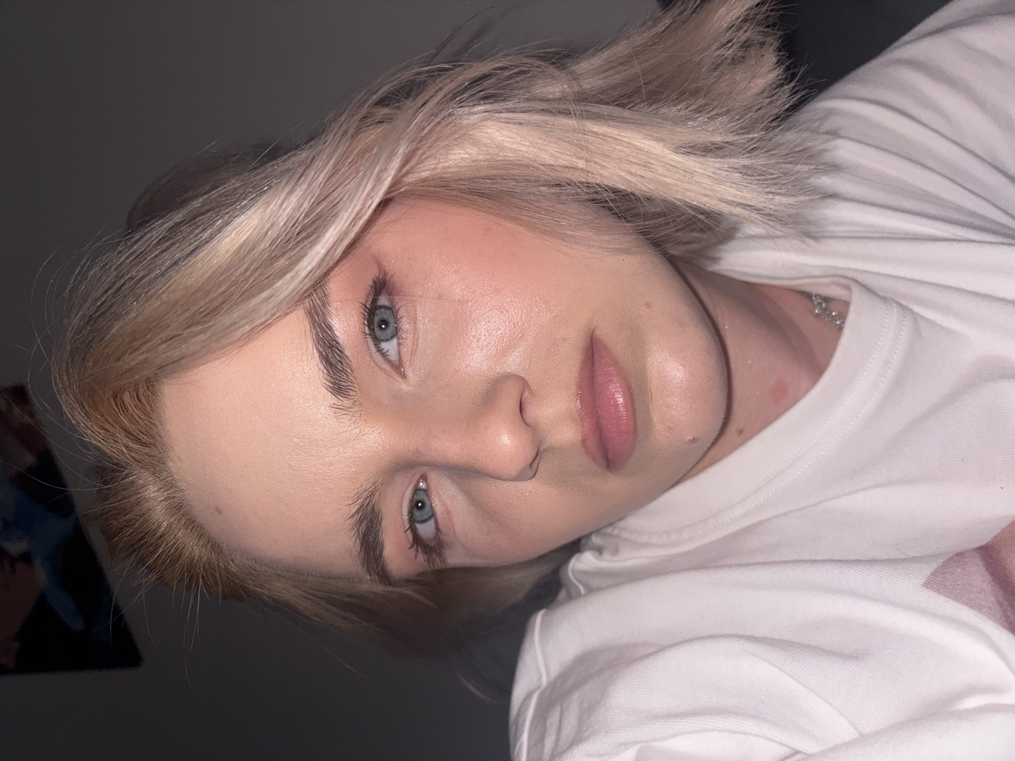 A close-up portrait captures a young individual with fair skin and light blue eyes, directly facing the camera with a neutral expression. The subject has short, platinum blonde hair with visible darker roots, styled in a bob. Subtle pink eye makeup enhances the eyes, complemented by mascara and well-defined eyebrows. The lips are glossy and naturally pink, and a small mole is present on the chin. The skin exhibits a dewy or slightly oily texture, particularly on the forehead and cheekbones. The individual is wearing a white crew-neck t-shirt, and a delicate silver chain necklace is visible around the neck. A small red mark or irritation is present on the upper chest/lower neck area. The background is dark and out of focus, suggesting an indoor setting with low lighting; a faint, blurry shape is discernible in the upper left. The lighting highlights the subject's face, creating soft shadows. This scene is captured indoors in the city of Ladozhskoe Ozero, Russia.