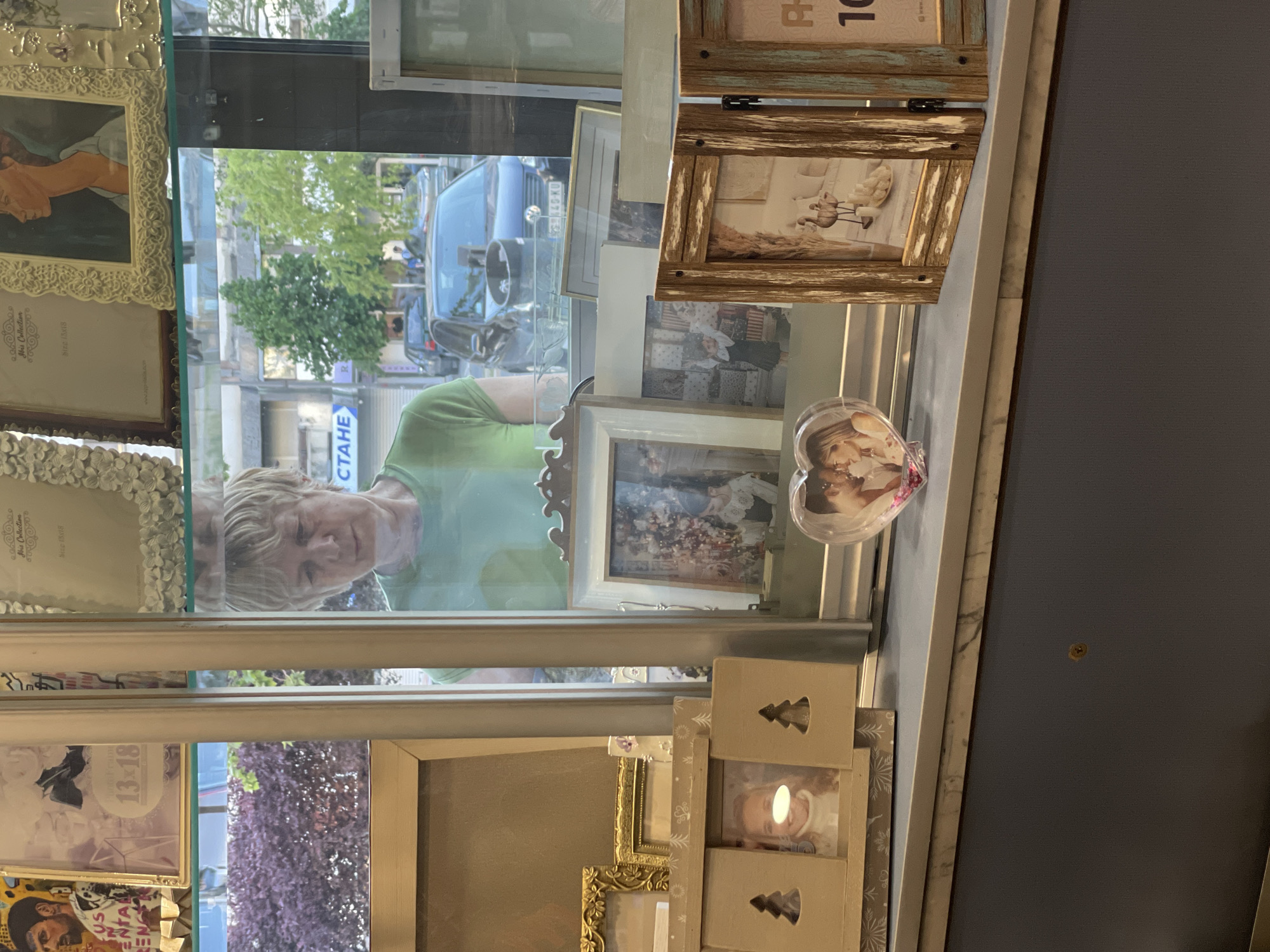 The image shows a reflection of a woman in the window of a shop. The shop appears to sell photo frames and other decorative items. The woman has short blonde hair and is wearing a green shirt. She seems to be looking at the shop display. Behind her, through the window, a street scene is visible with cars and trees. A sign with Cyrillic letters (СТАНЕ) can be seen, suggesting the image was taken in a country that uses the Cyrillic alphabet. Based on the sign and the context, it is possible that the location is in the city of Novi Sad, Serbia.