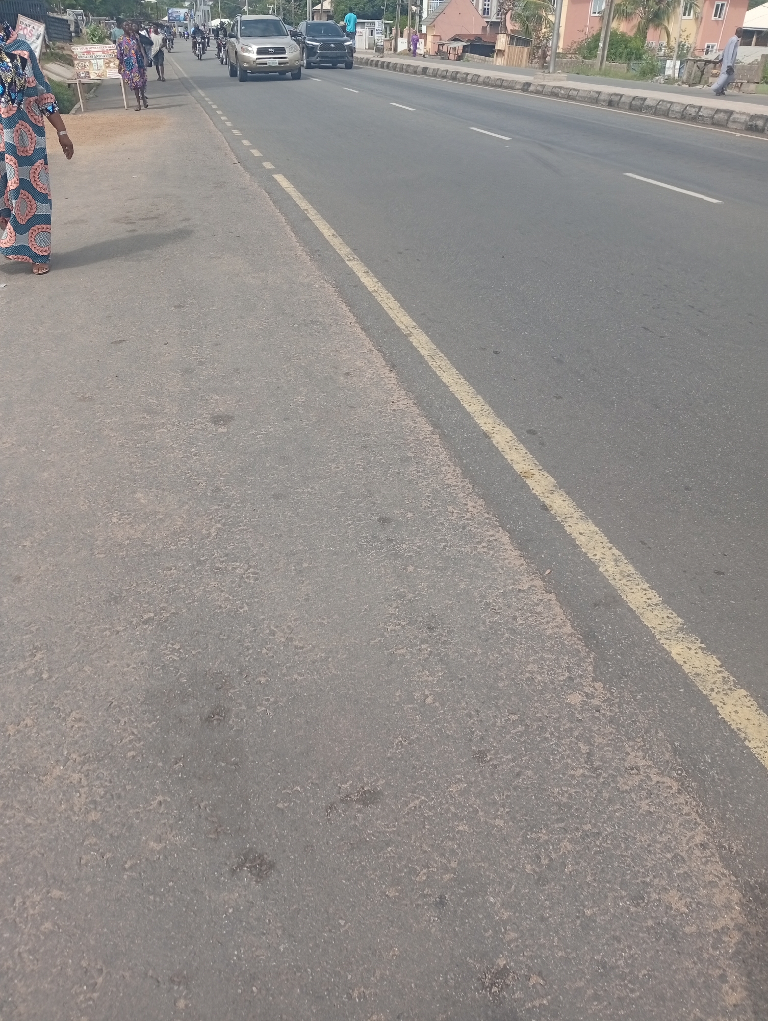 The image depicts a street scene in Adankolo, Nigeria. The street is paved and has a yellow line marking the edge of the road. In the distance, cars and motorcycles can be seen driving on the road. On the left side of the road, a woman is walking along the sidewalk. Other individuals and buildings are visible in the background.