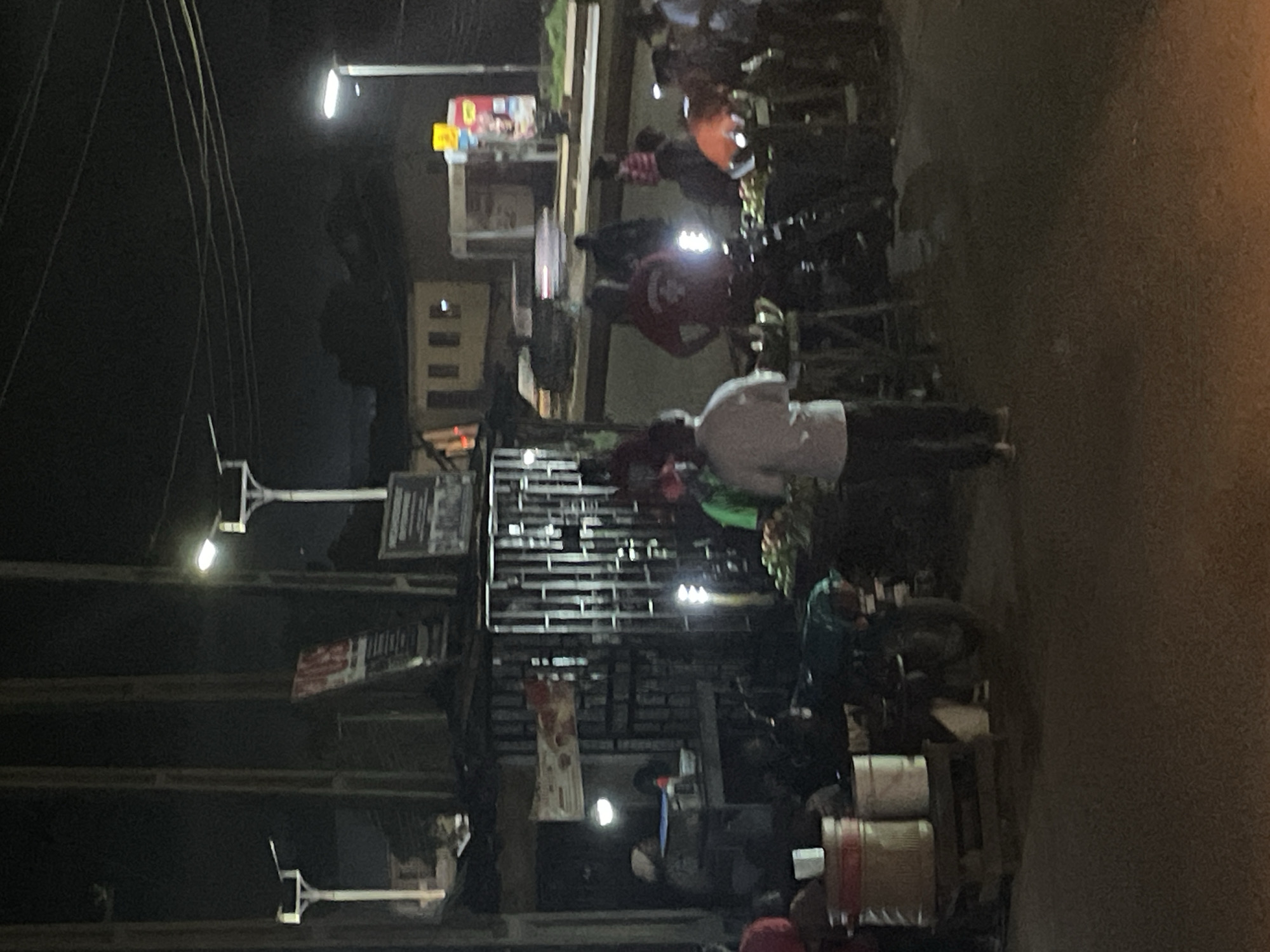 A group of people are gathered at an outdoor market at night in an unknown city. Several stalls are visible, with some selling produce. There are also streetlights illuminating the scene. The image is blurry due to low light conditions and possibly motion.