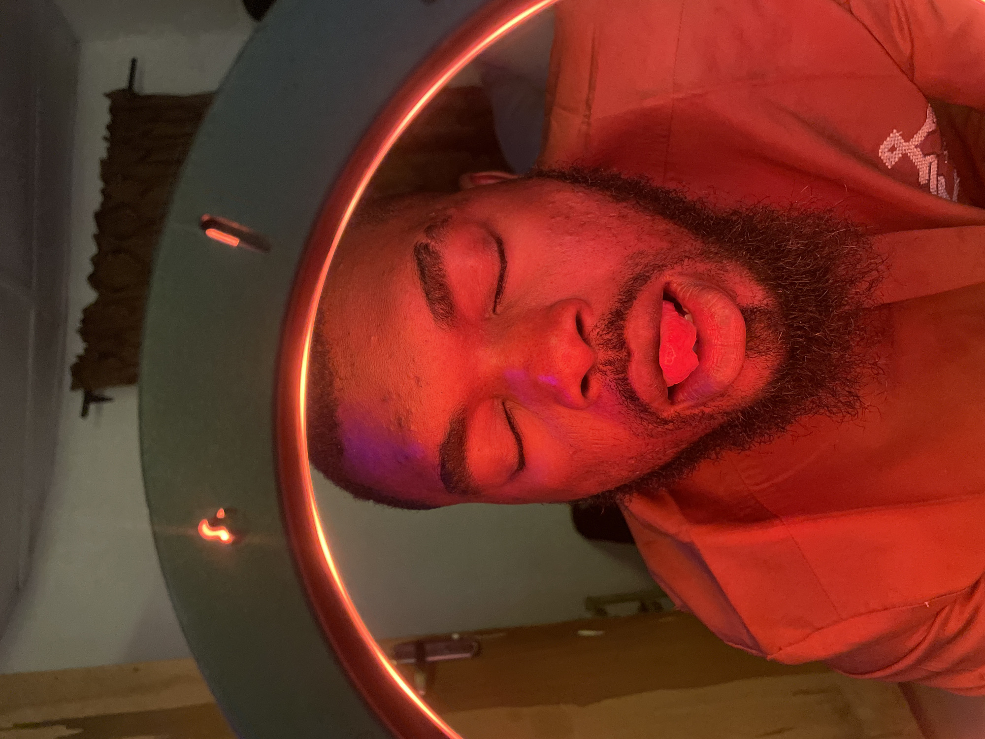 The image shows a young Black man posing, rotated 90 degrees clockwise from a typical upright orientation. He is illuminated primarily by a strong, circular red light fixture, creating a dramatic and atmospheric effect. The man has a beard and is depicted with his eyes closed and his tongue slightly extended, giving the impression of a playful or artistic pose. He is wearing a reddish-orange short-sleeved shirt with a white, stylized emblem on the upper right chest area.
His head is positioned near the edge of a light-colored, possibly pale green or teal, table or counter surface. On this surface, a distinct glowing red Apple logo is visible. In the background, a textured wall, possibly adorned with rough-hewn stone or a similar decorative panel, can be seen. The overall setting appears to be indoors, likely a dimly lit room, with the red light being the primary illumination source, suggesting a nighttime scene. There's a faint hint of purple or blue light on the left side of his face. The location context is Jos, Nigeria.